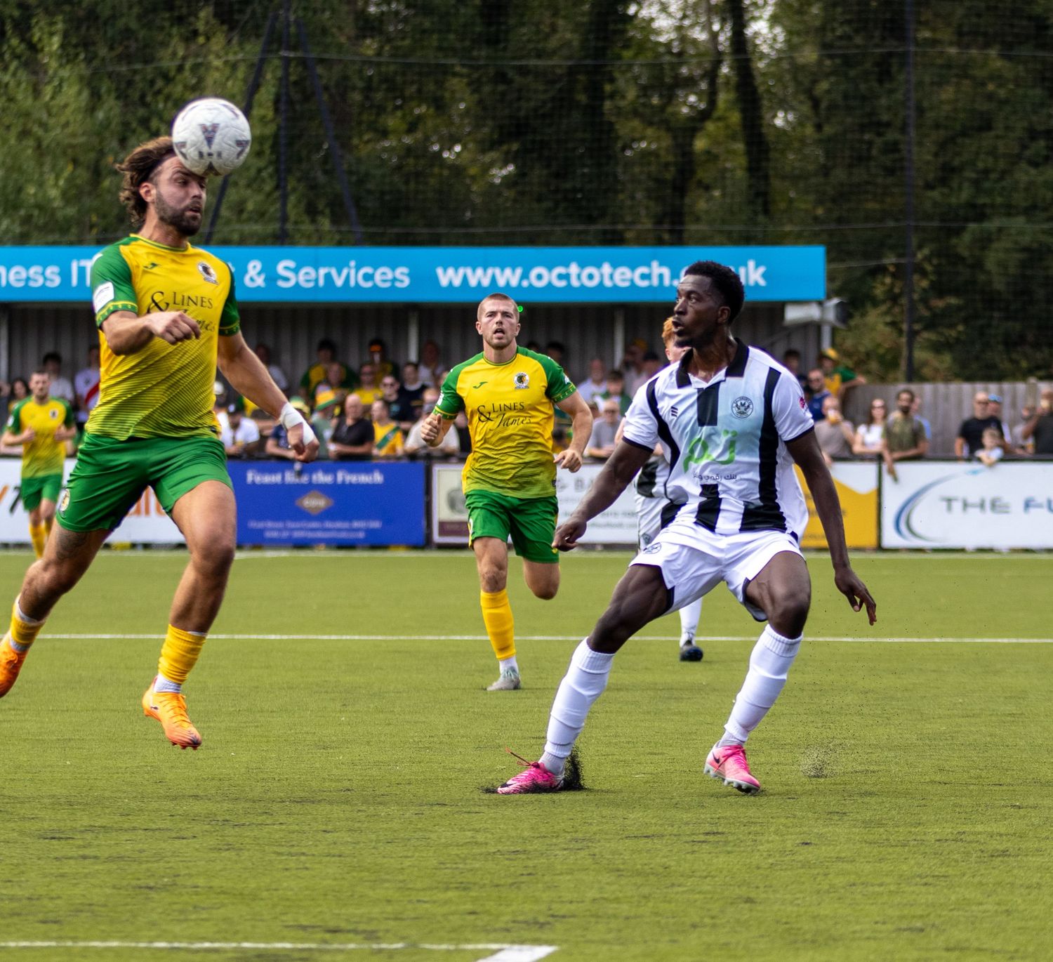 Footballers in yellow-green and black-white kits contest aerial ball during outdoor football match.