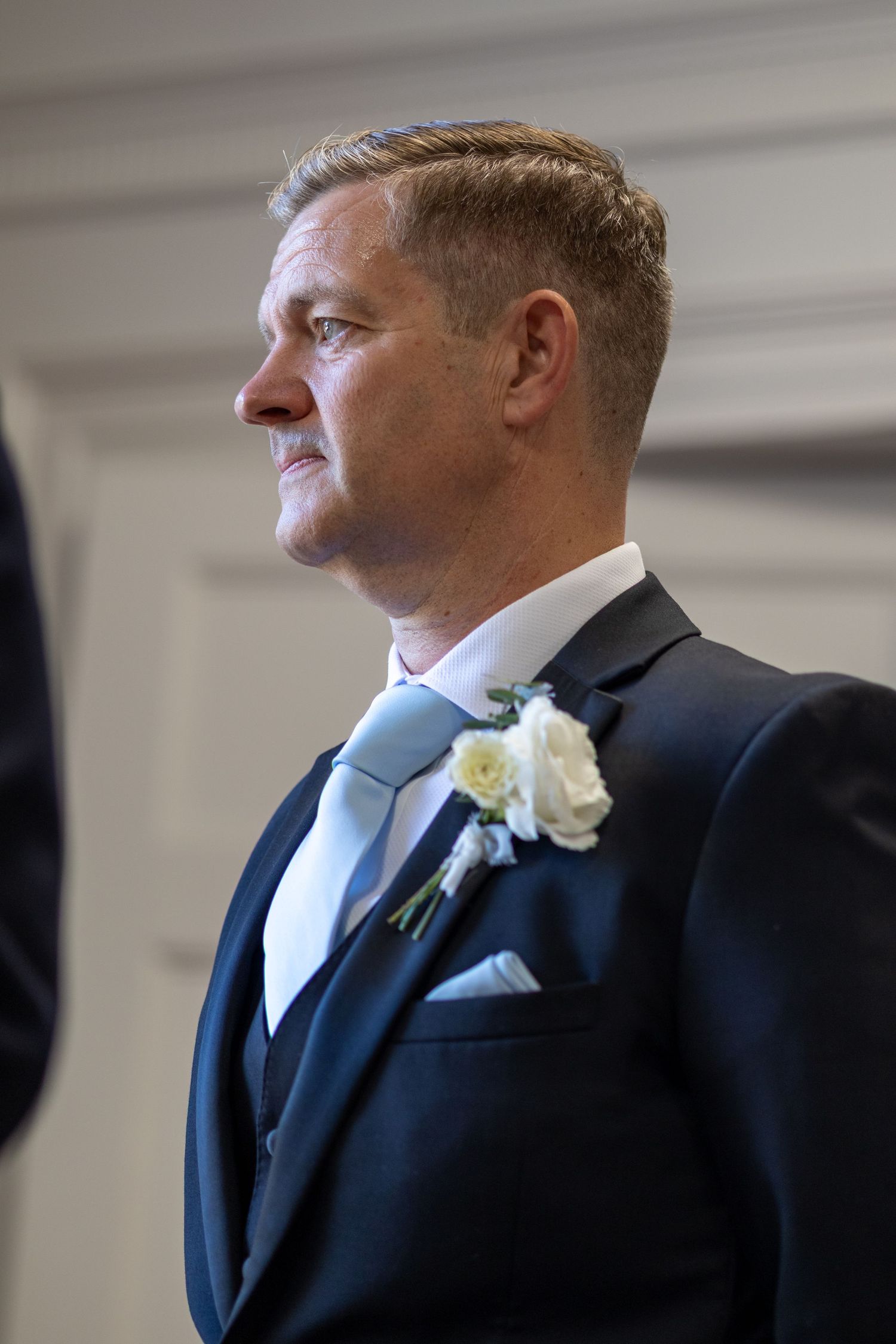 Elegant groom in navy suit with white rose boutonniere and light blue tie at wedding ceremony.