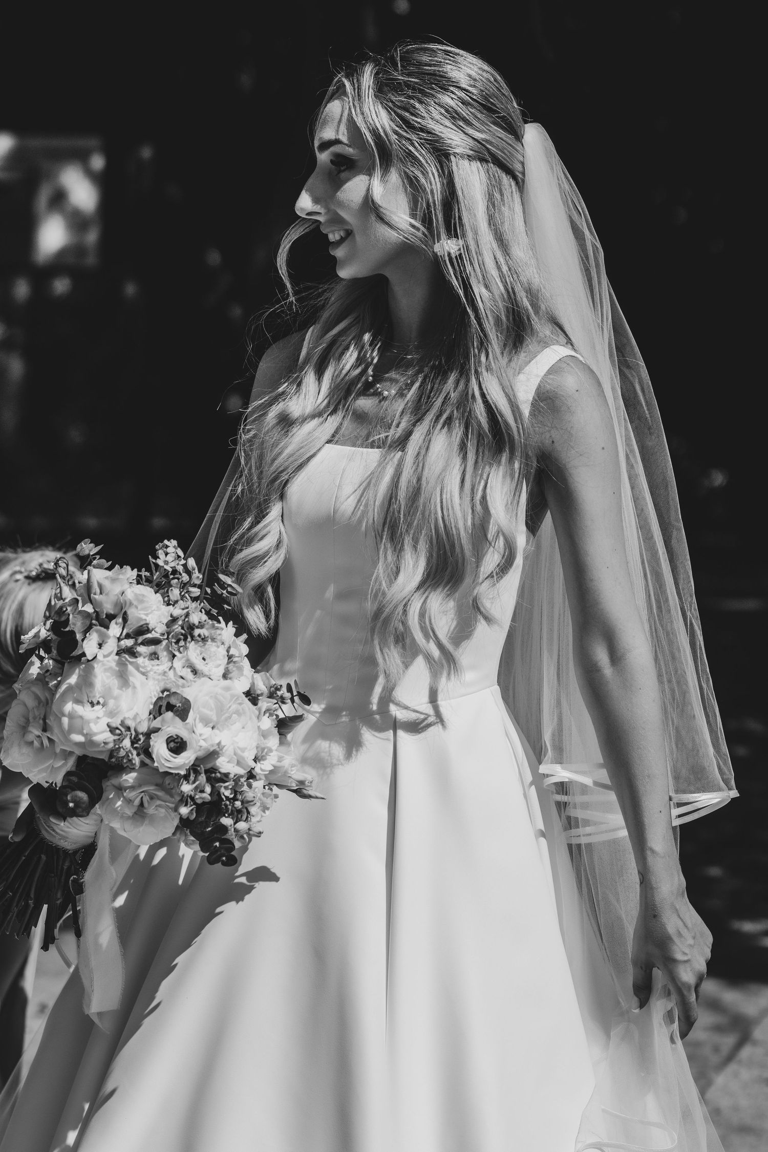 Black and white portrait of a radiant bride in a white gown holding a floral bouquet with a veil.
