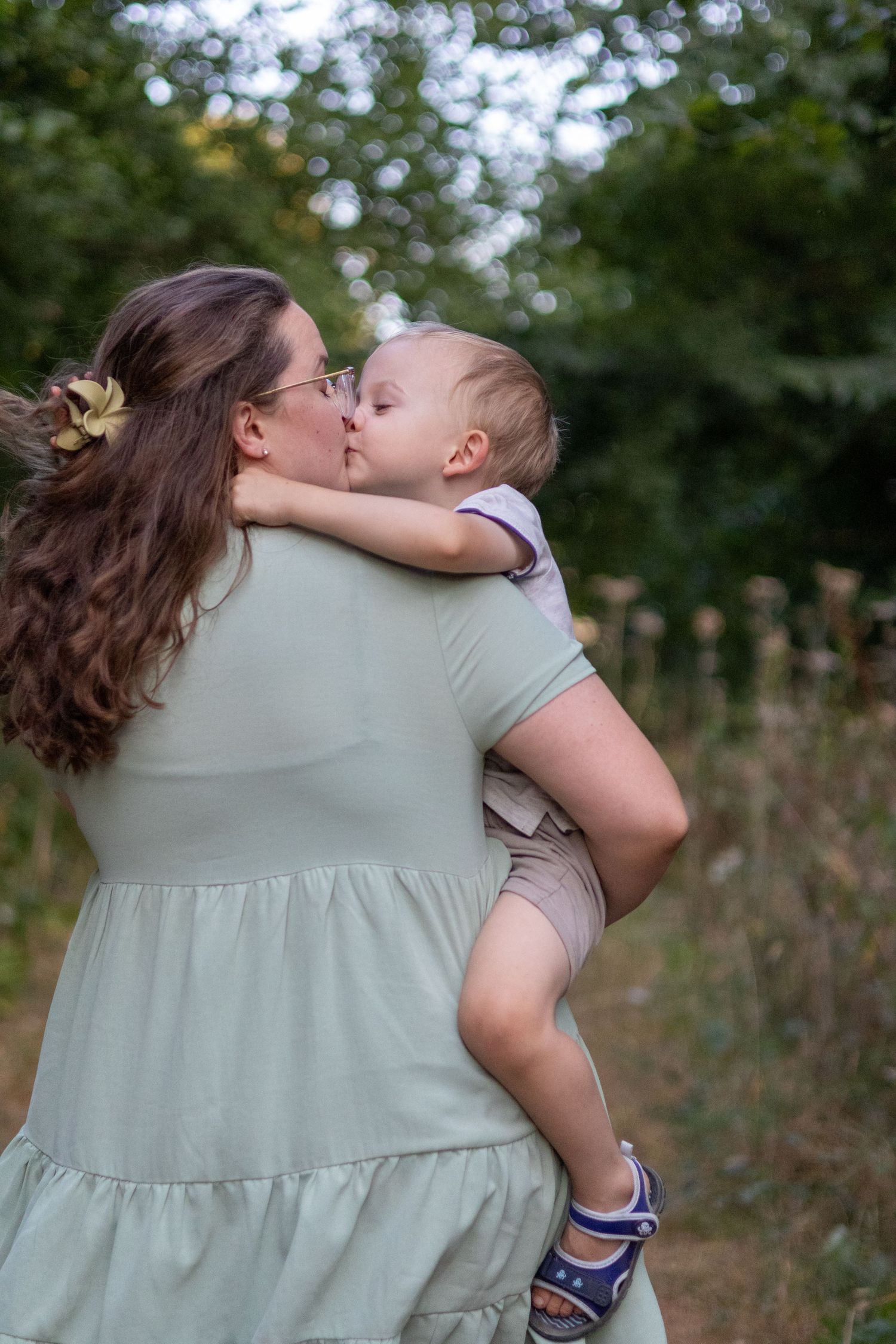 Mother in sage dress kissing her baby on a peaceful tree-lined garden path.