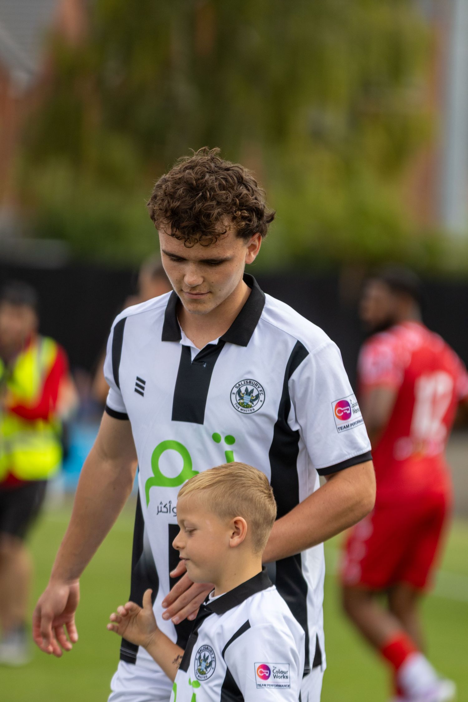 A young footballer in a black and white kit walks with a mascot child on the pitch.