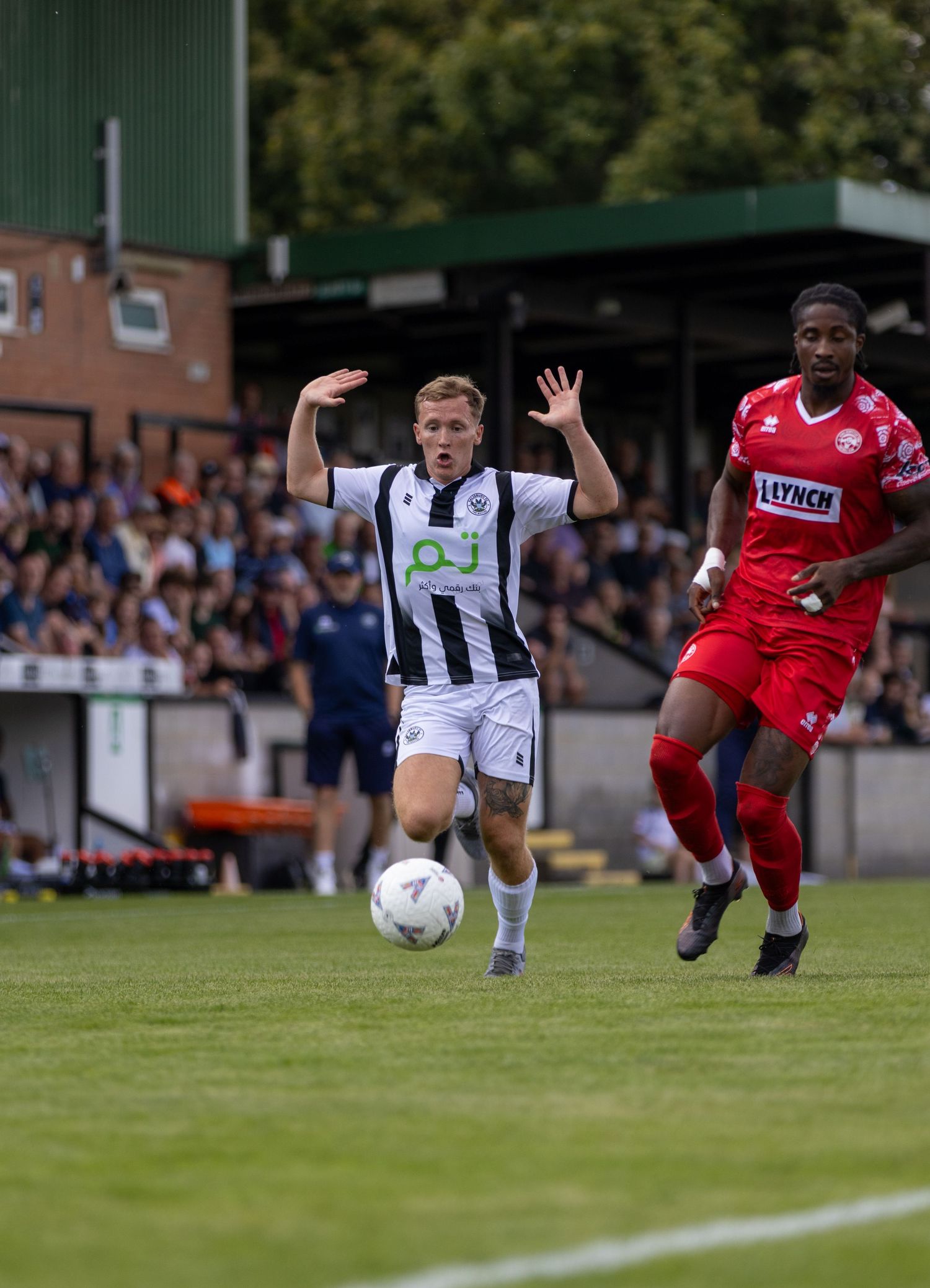 Blonde footballer in black and white stripes chases the ball ahead of a red-kitted opponent on a sunny matchday.