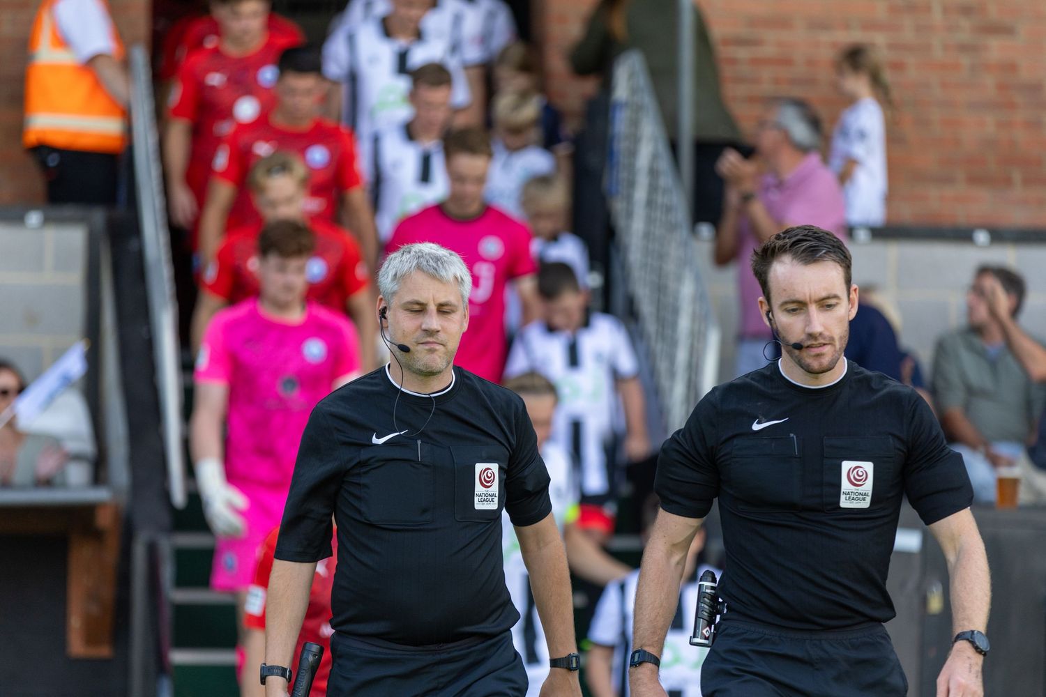 Two football referees in black uniforms walking onto the pitch before a match, with players visible in the background.