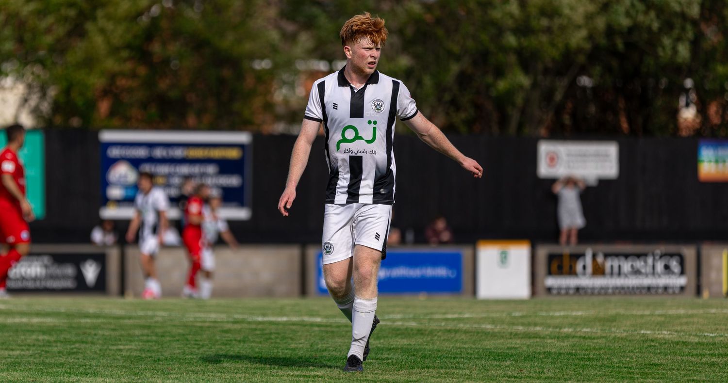 A young male soccer player in black and white striped kit jogging on a sunny outdoor football pitch during a match.