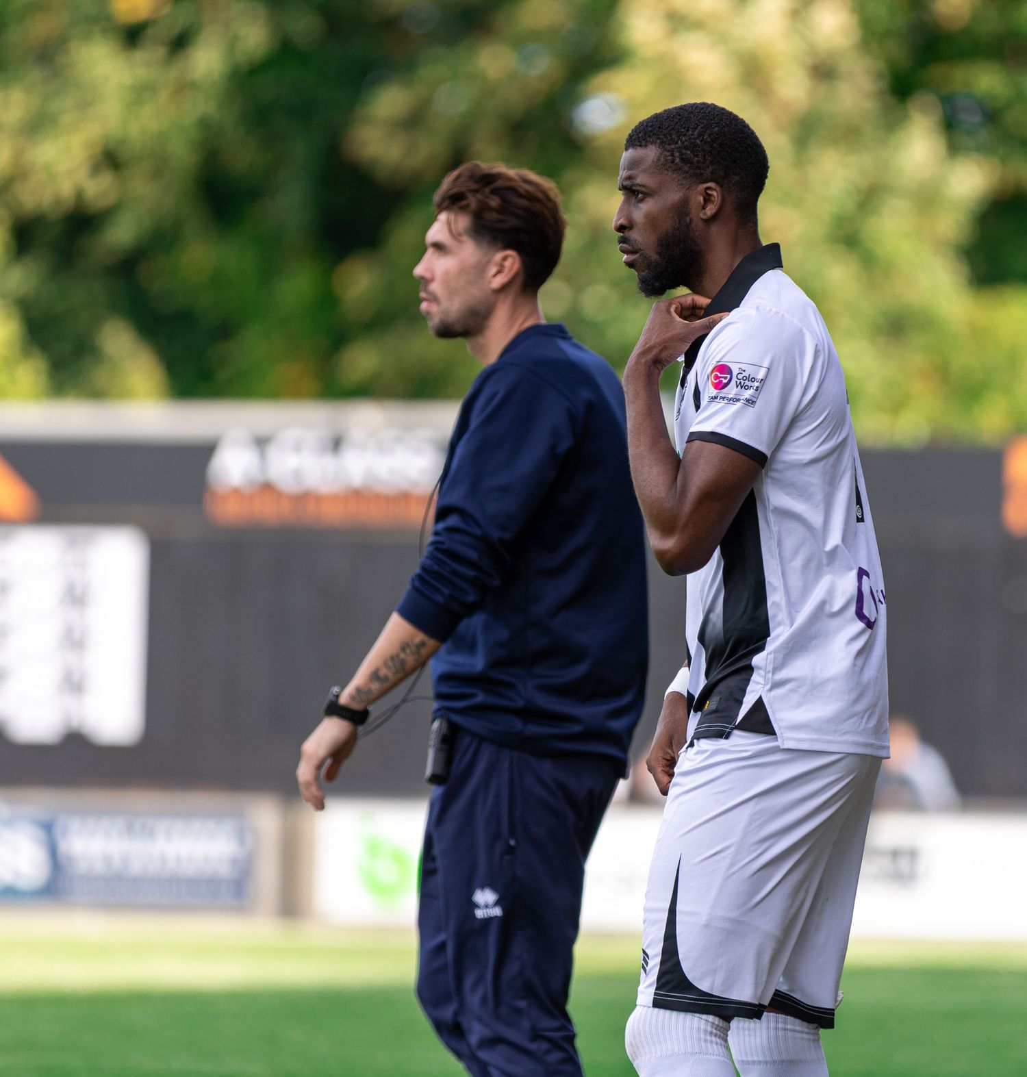 Two soccer players in white uniforms stand on a field during a match, with a coach in navy blue nearby.