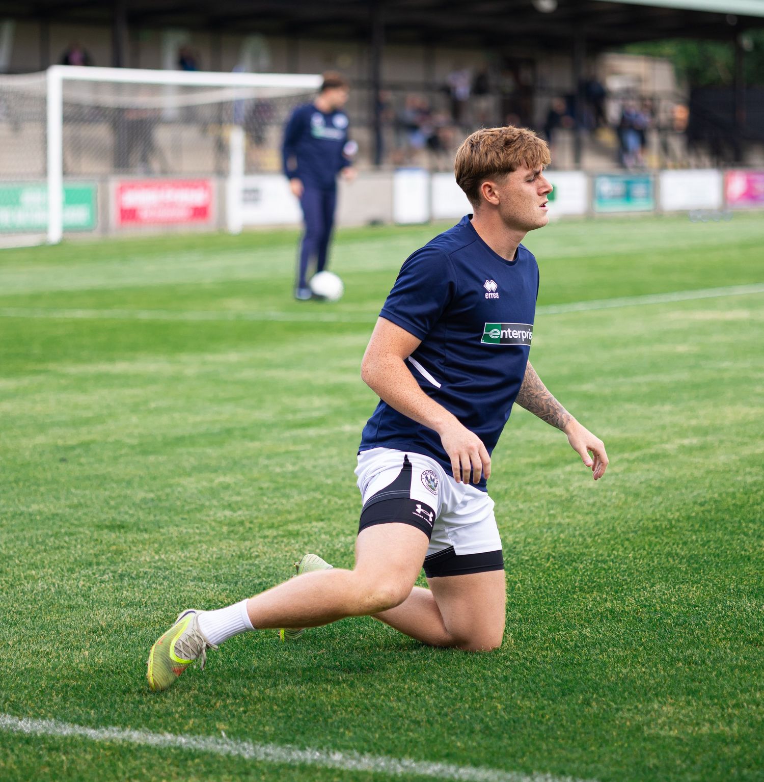 A young footballer in navy kit stretches on a green pitch during pre-match warm-up at a football stadium.
