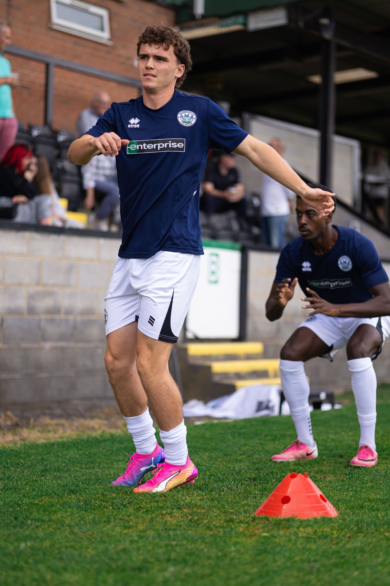 Two football players in navy blue kits warm up on a grass pitch, one stretching near orange cones during pre-match preparation.