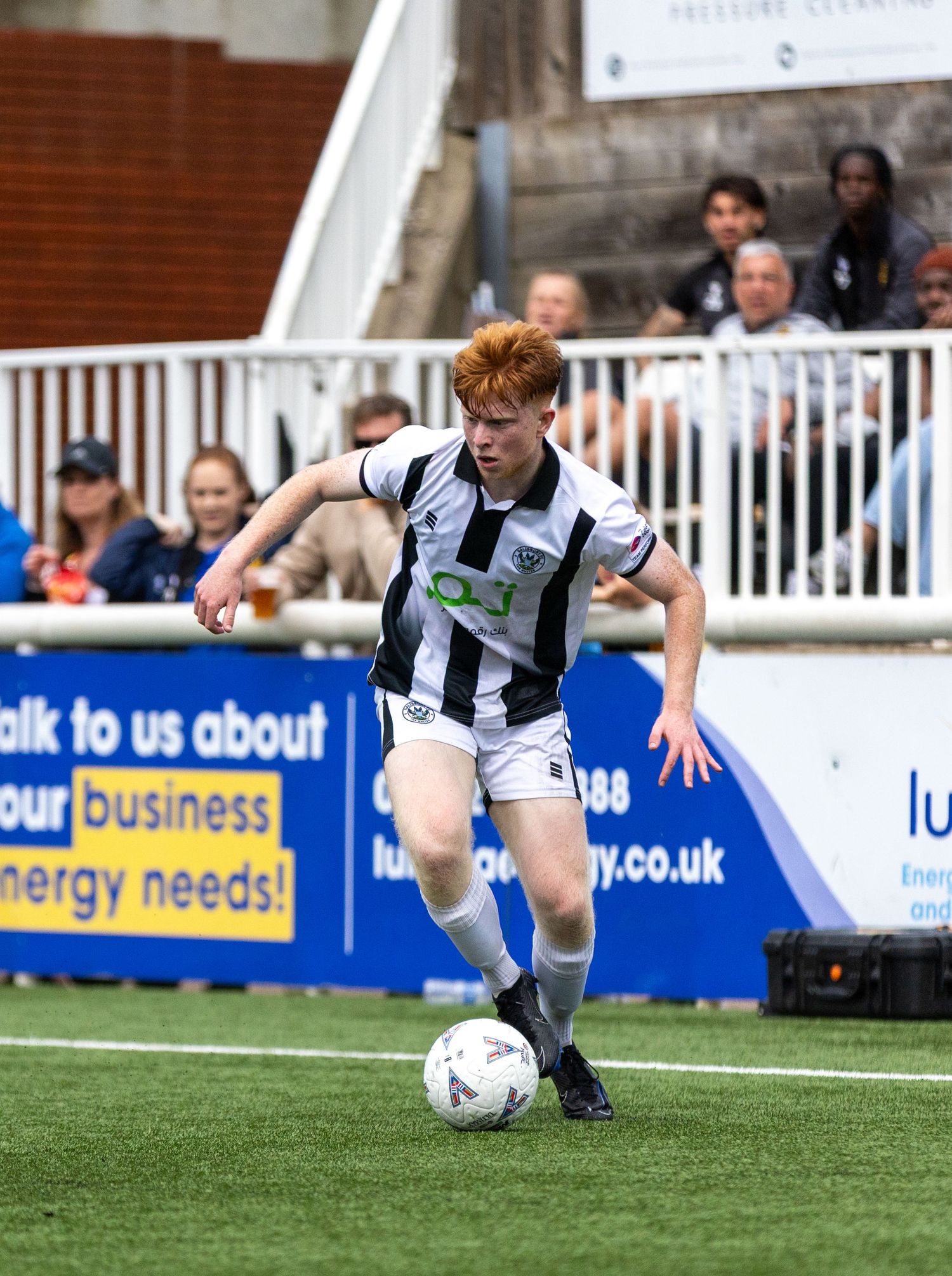 Young red-haired footballer in black and white kit dribbling a ball during a competitive match.