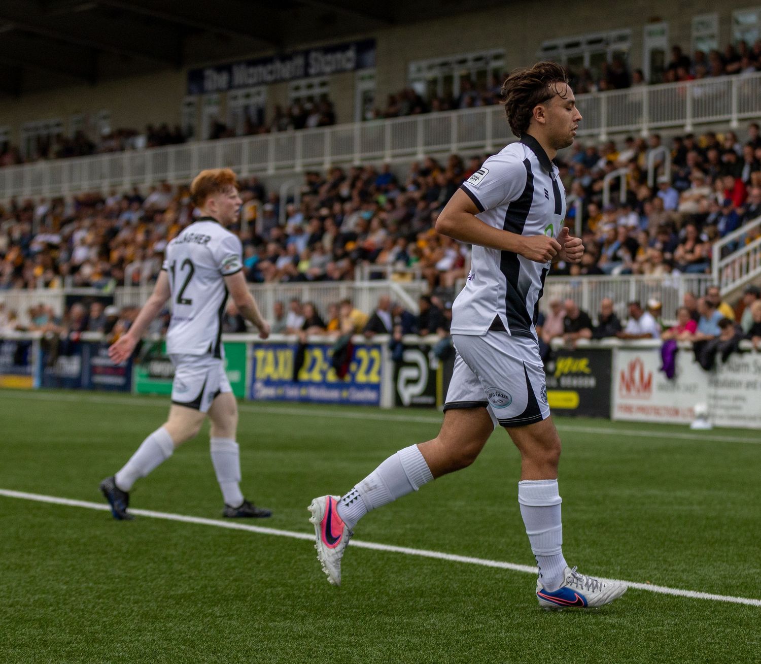 Two soccer players in black and white uniforms jogging on a football pitch during a match with fans in the stands.