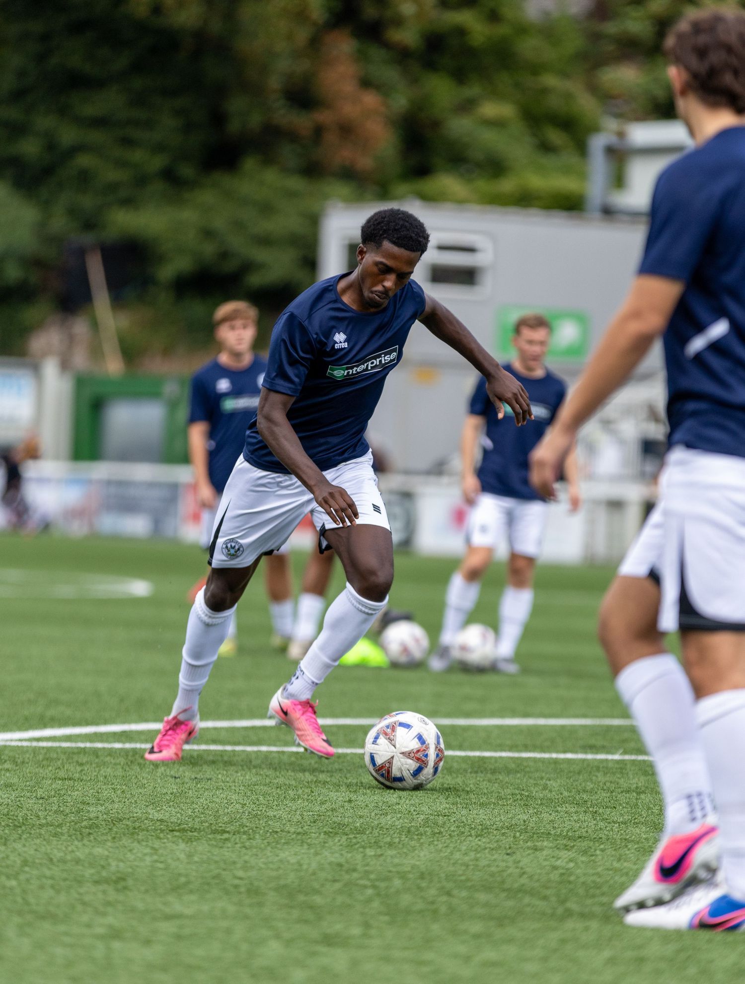 Soccer player in navy blue kit dribbling ball on green turf during a match with teammates in background.
