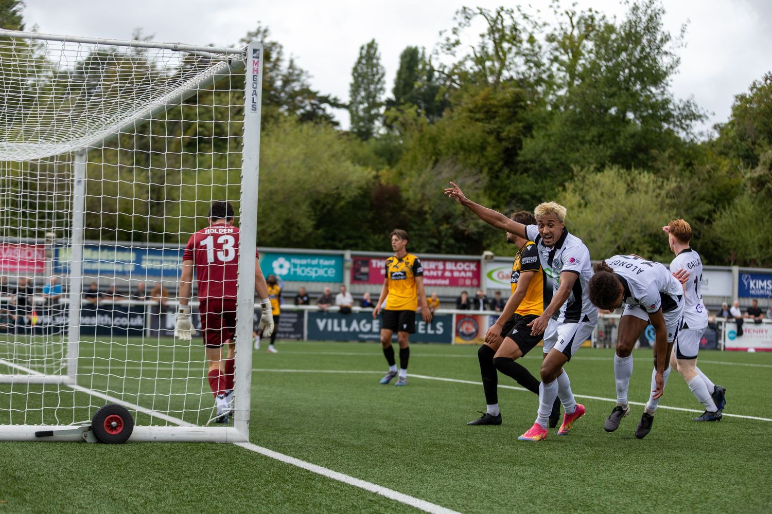 Soccer players compete near the goal during a non-league football match on an artificial turf pitch.