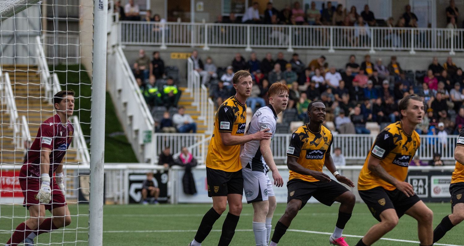 Soccer players compete near the goal during a match, with fans watching from the stands in the background.