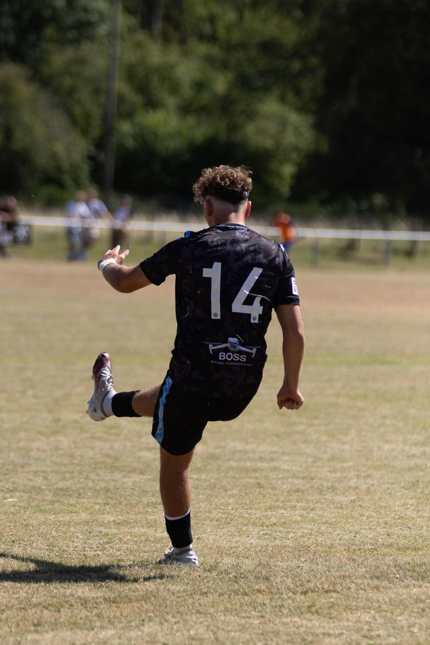 Football player number 14 in black kit kicking ball during sunny outdoor match.