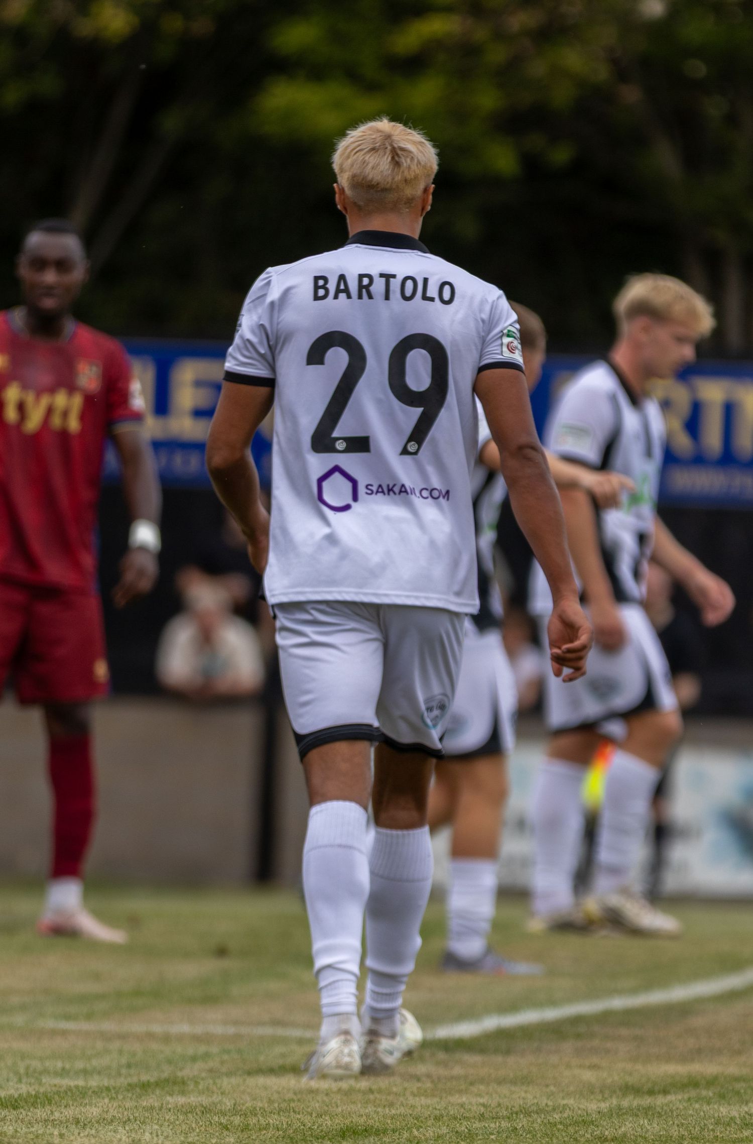 A footballer wearing white number 29 Bartolo jersey with Saka.com sponsor walks on pitch.