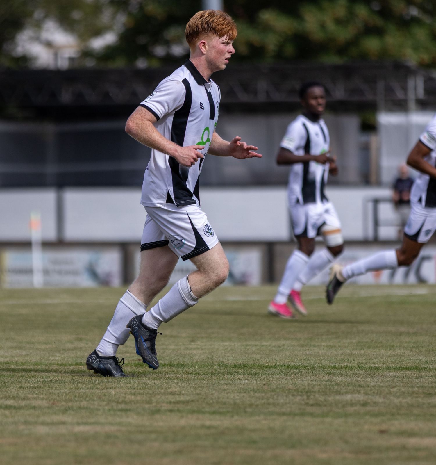 Young red-haired soccer player in white and black kit sprints across green pitch during a match.