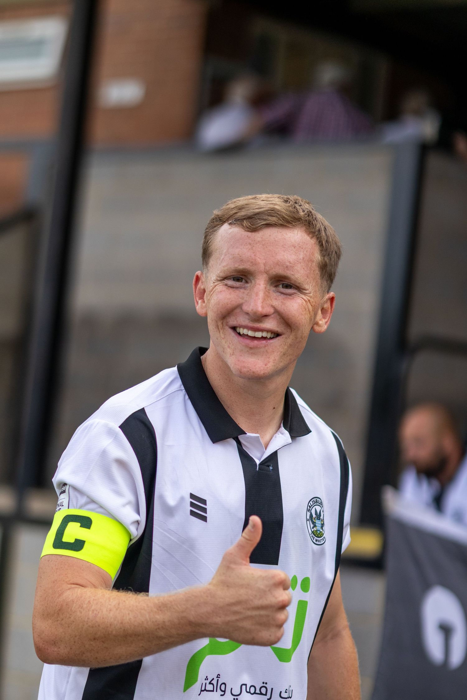 A smiling male soccer player in a white and black jersey gives a thumbs up wearing a captain's armband.