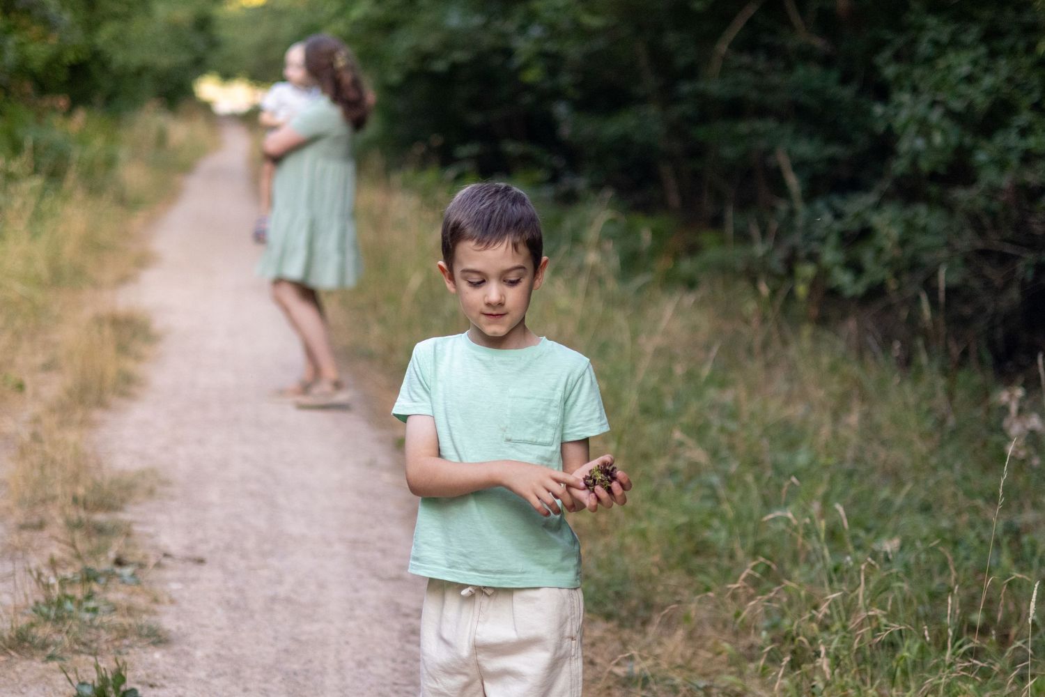 A young boy in a mint green shirt examines something in his hands on a dirt path in nature.