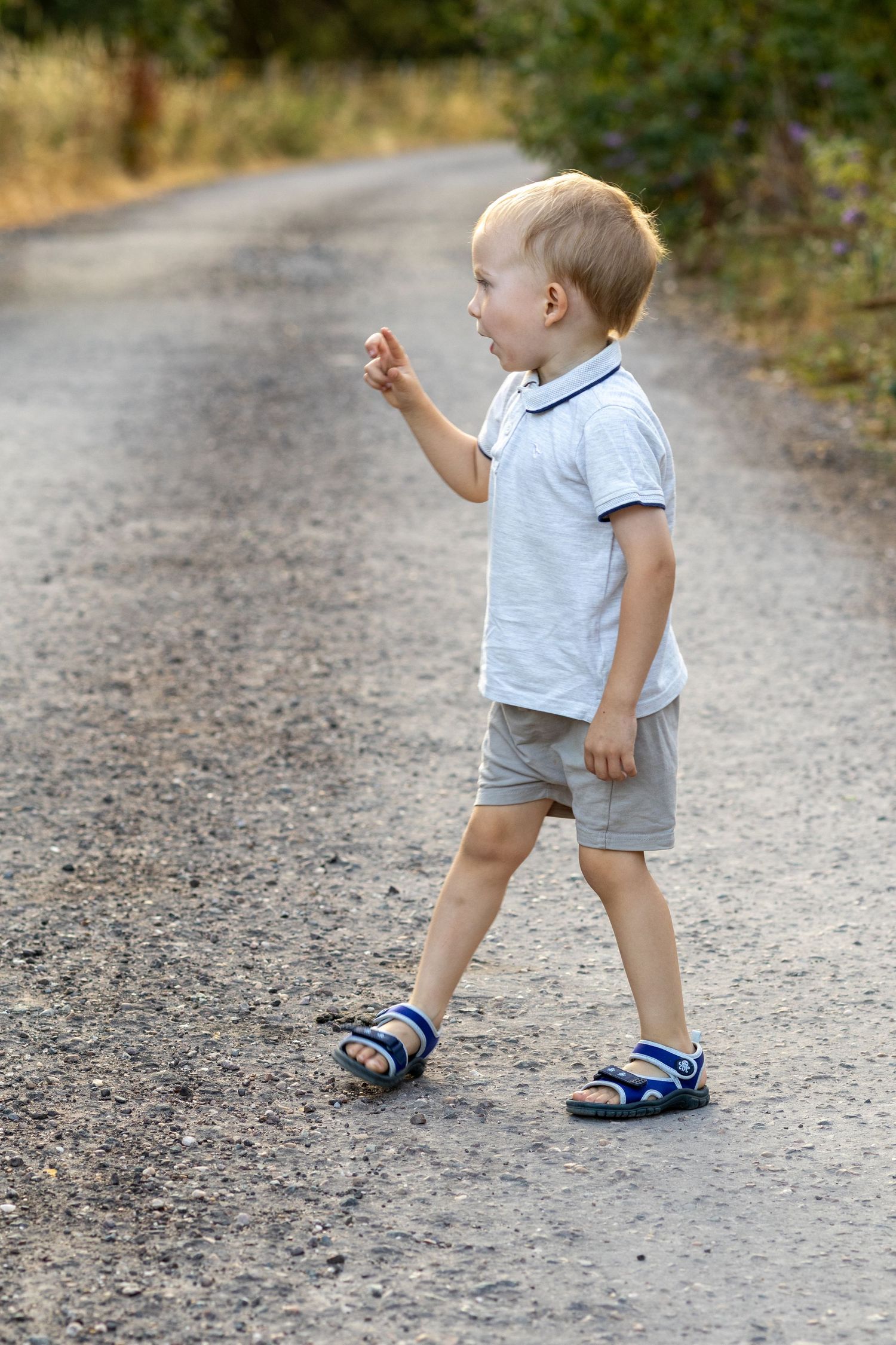 Toddler boy in white polo shirt and gray shorts walking on a gravel road outdoors.