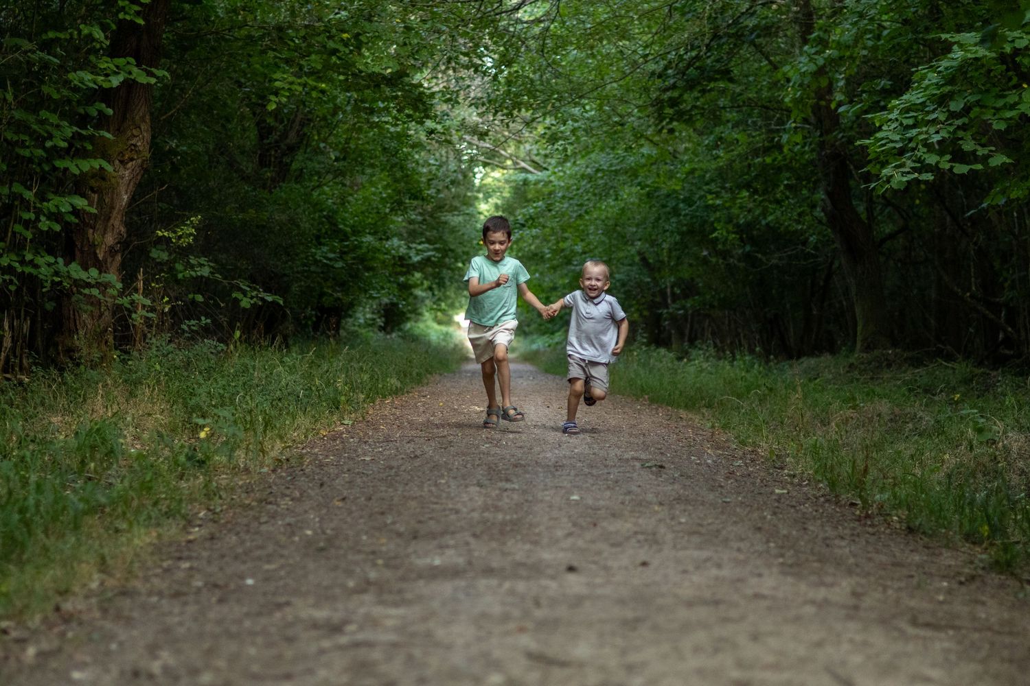 Two young children holding hands running joyfully down a tree-lined forest path.
