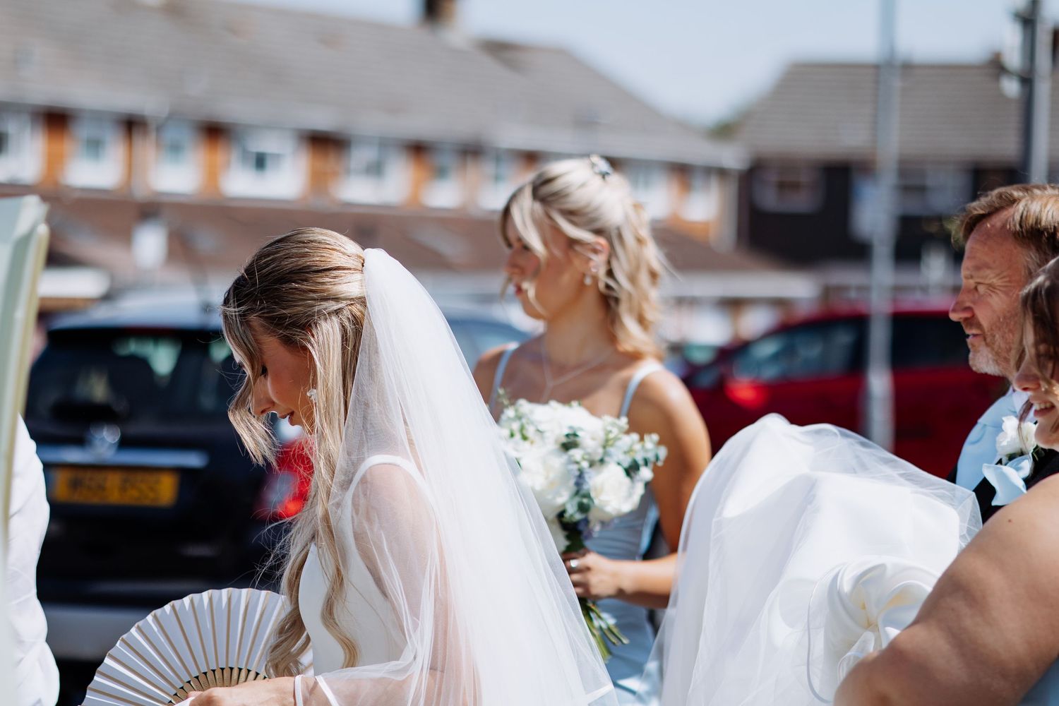 Bride in white wedding dress and veil arriving at church with wedding party on a sunny day.