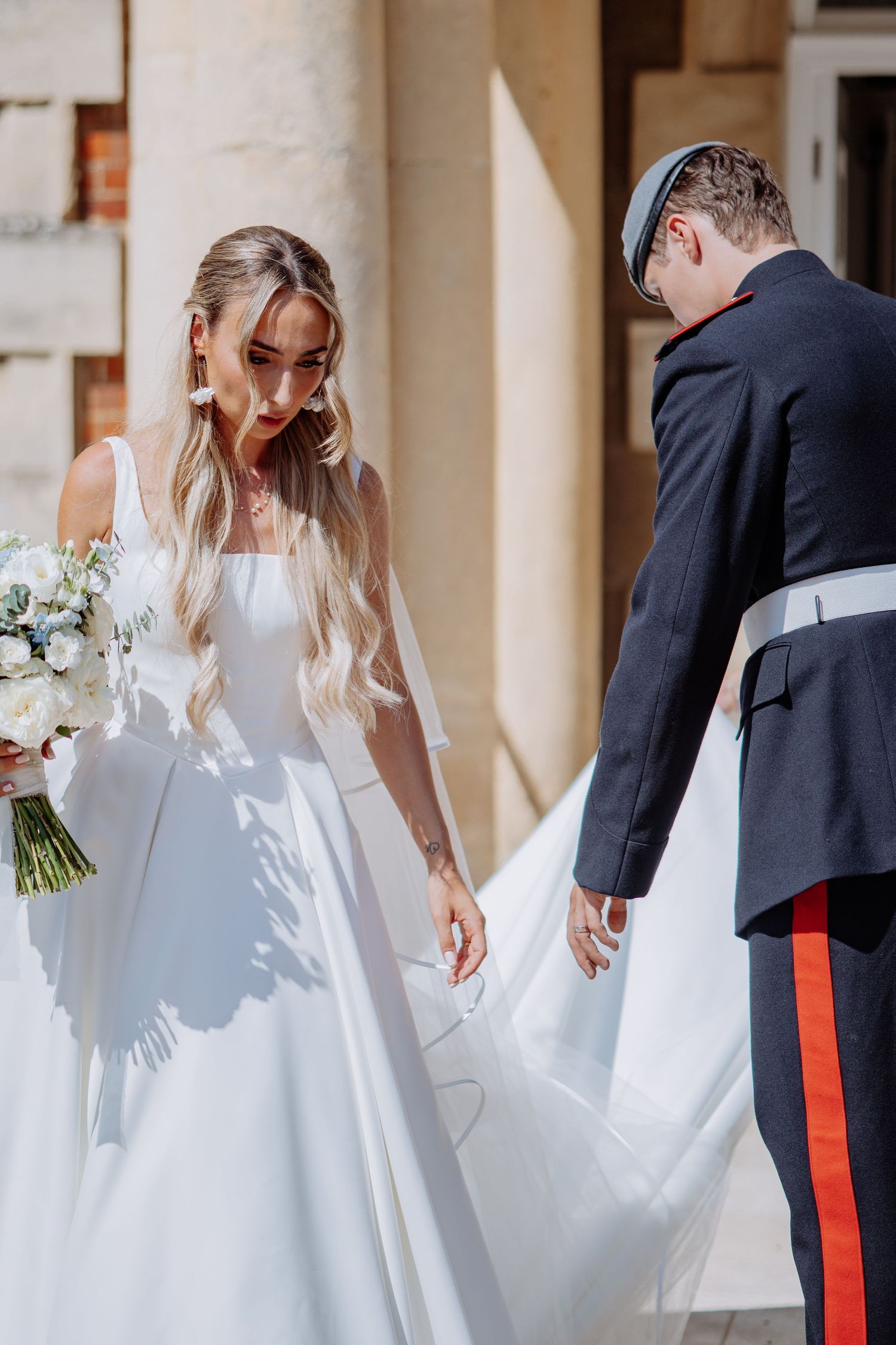 Military groom helps bride with wedding dress train in sunny courtyard.