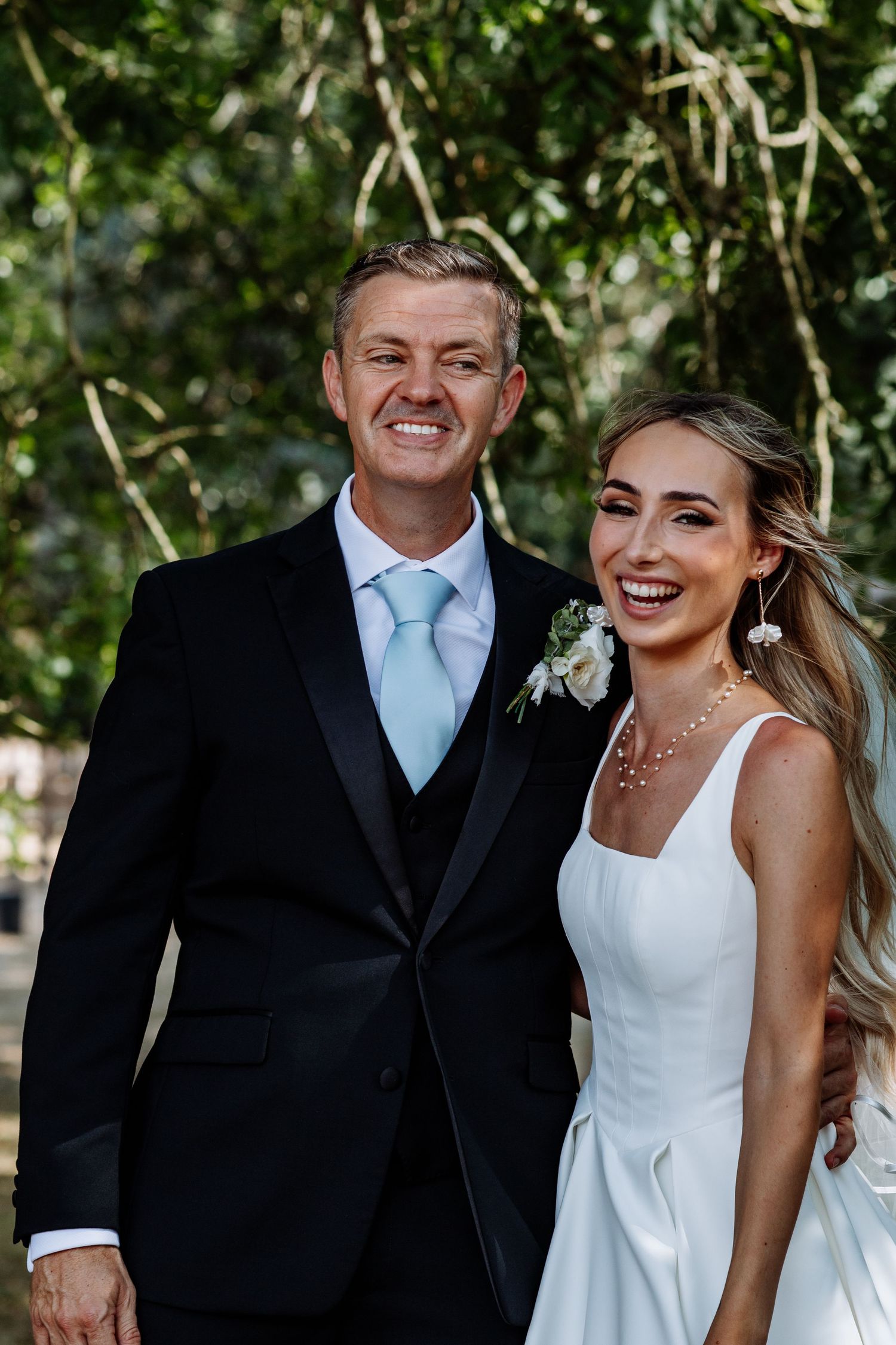 Happy couple in wedding attire smiling together outdoors with lush green trees in background.