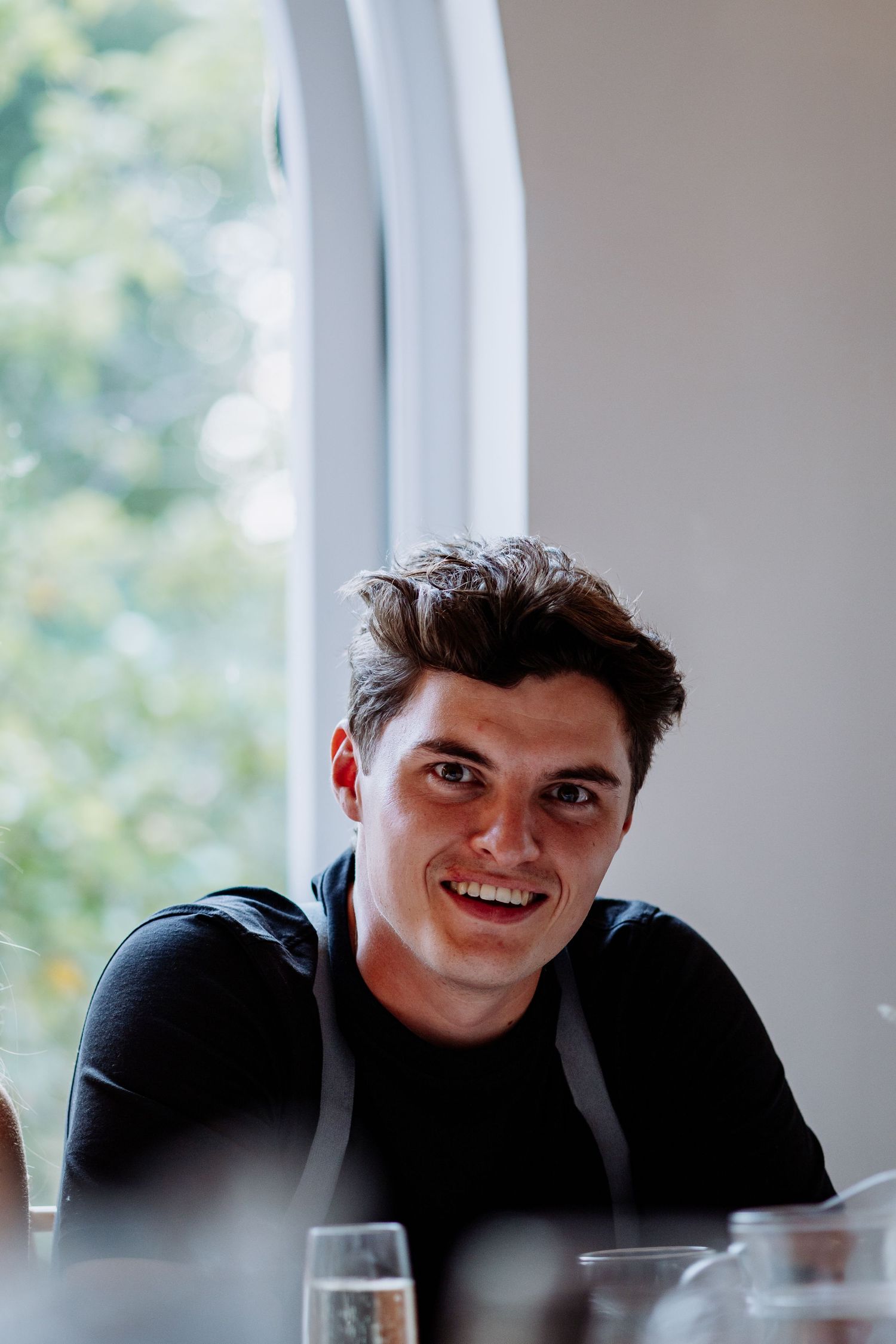 Smiling young man with dark hair in black shirt seated at wedding reception table with champagne glasses.