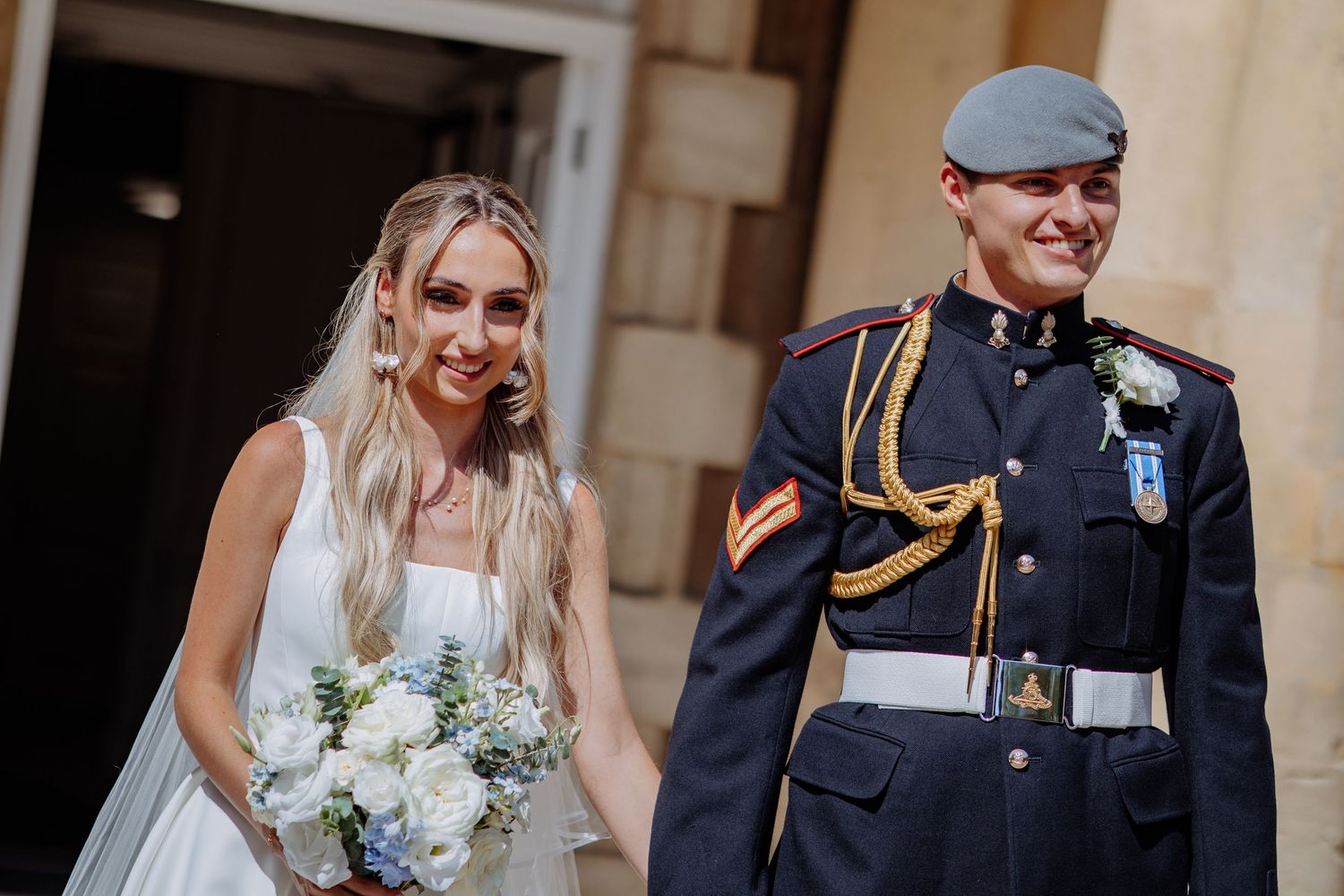 Bride in white sleeveless wedding dress with bouquet walks alongside groom in RAF dress uniform at elegant venue.