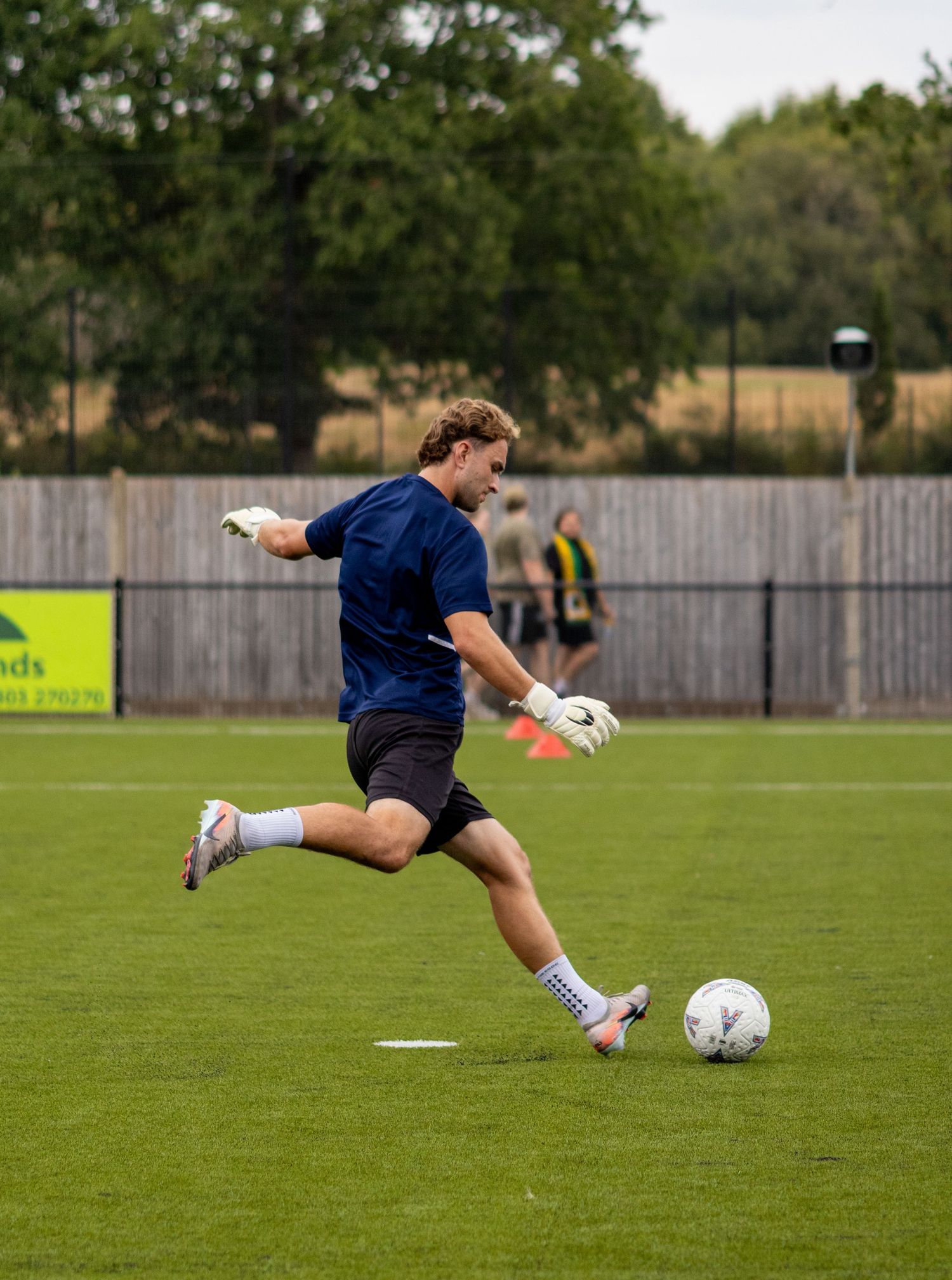 Goalkeeper in navy kit and white gloves taking a powerful kick during pre-match warmup on grass pitch.
