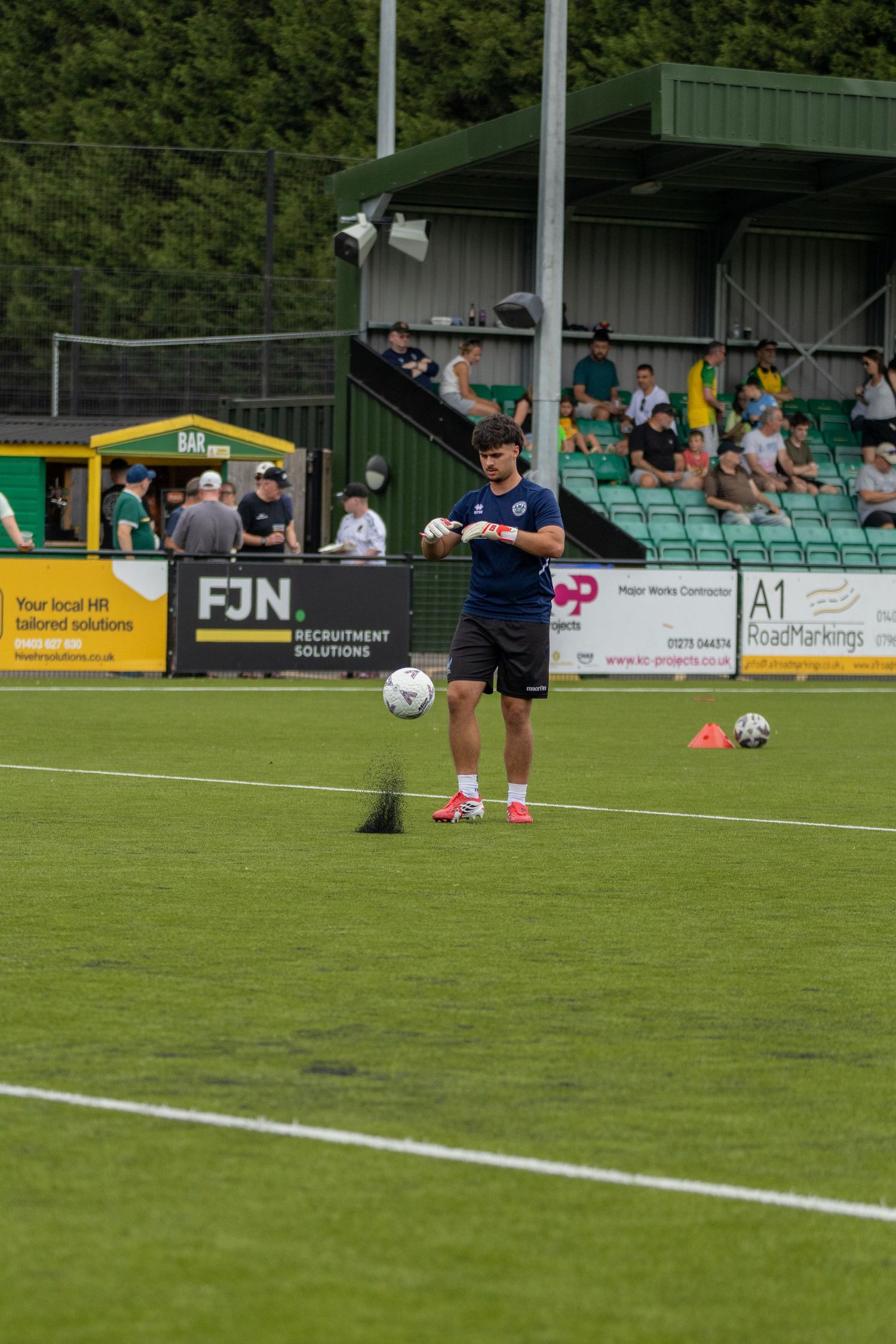 Football coach in navy kit holding ball during pre-match warmup at green stadium with FJN Recruitment Solutions signage.