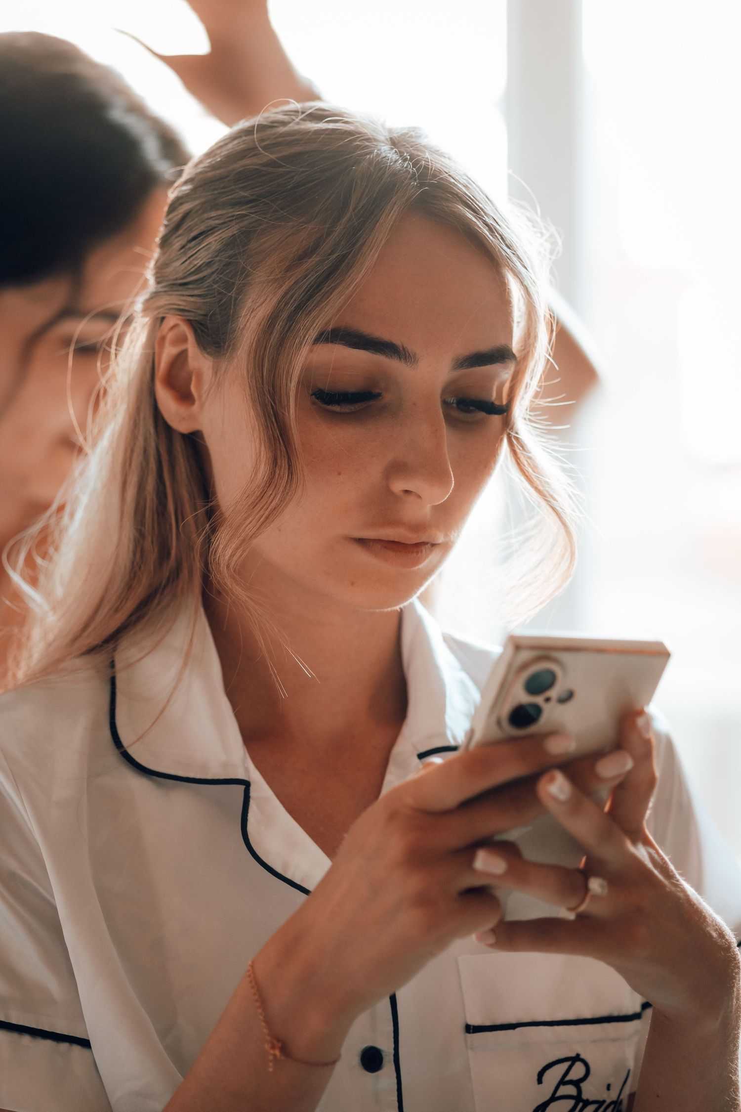 Bride in white pajamas looking at her phone while getting ready on her wedding day.