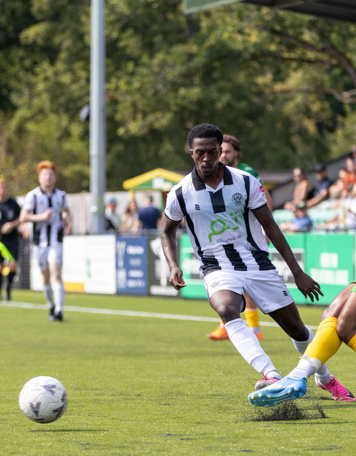 A footballer in black and white striped kit dribbles a ball on a sunny green pitch during a competitive match.
