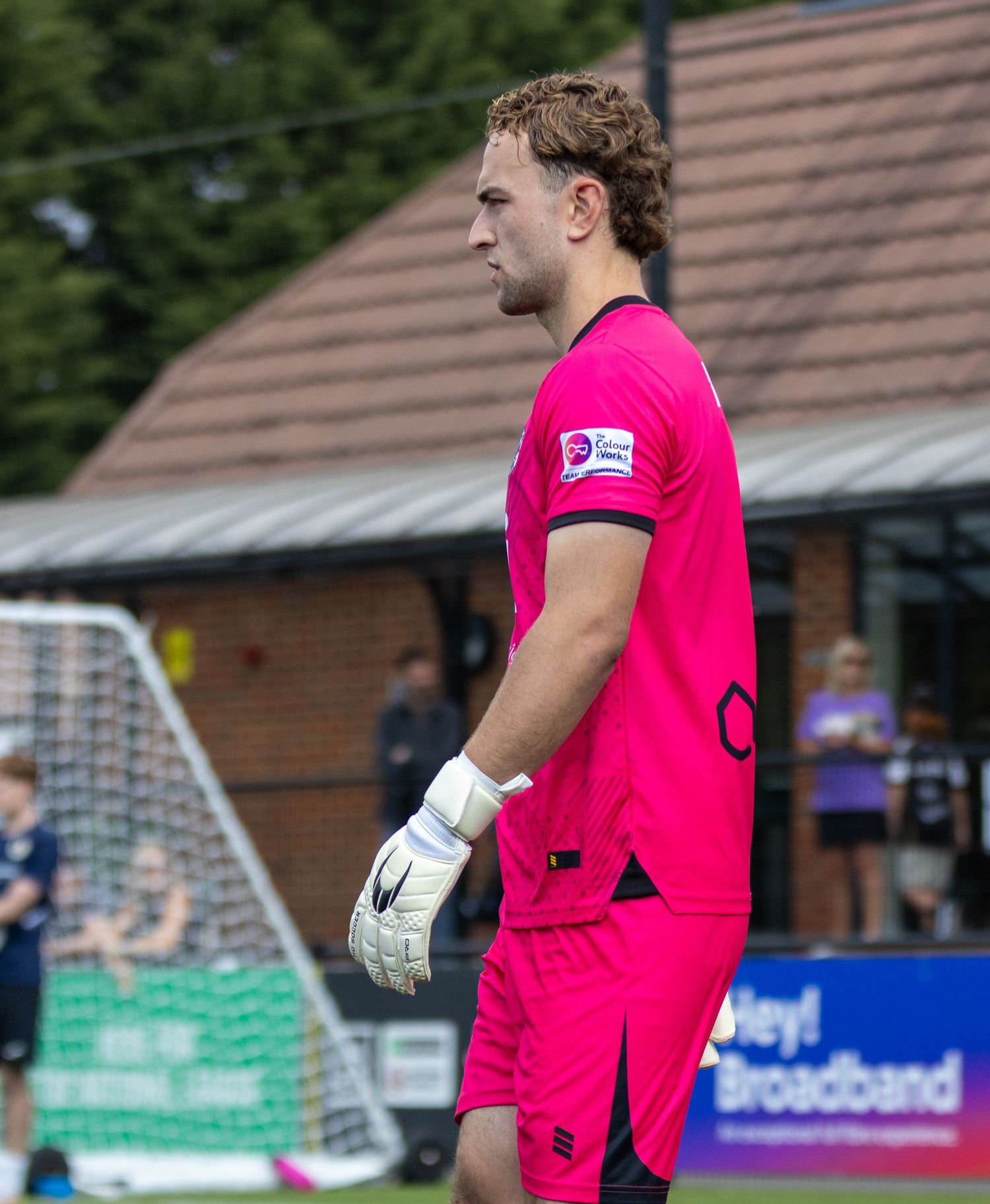 A goalkeeper in a bright pink kit stands on the pitch during a non-league football match on a sunny day.