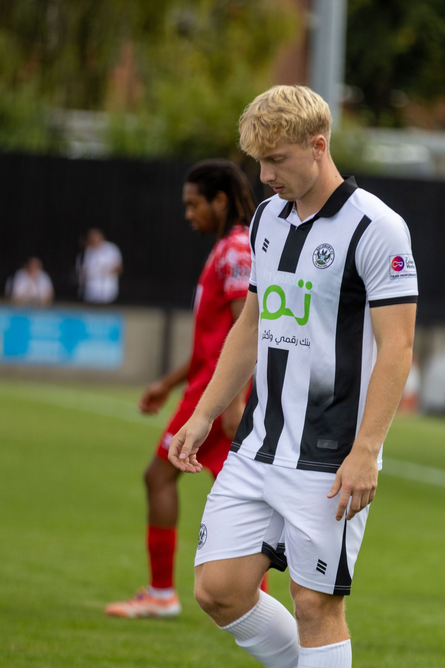 A young blonde footballer in a black and white striped kit walks on the pitch during a football match.
