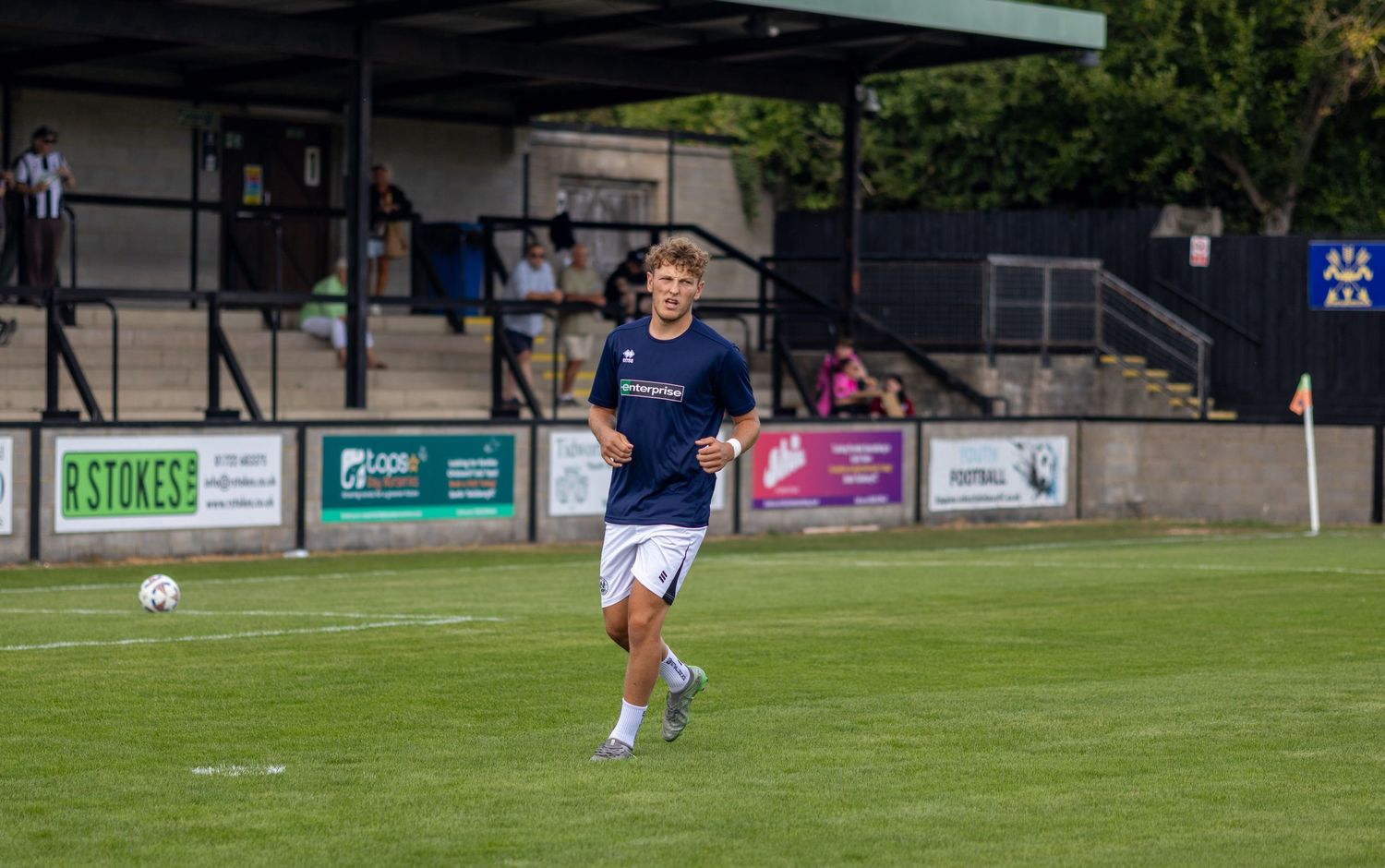 Young soccer player in navy blue kit jogging across a green pitch during pre-match warmup at a stadium.