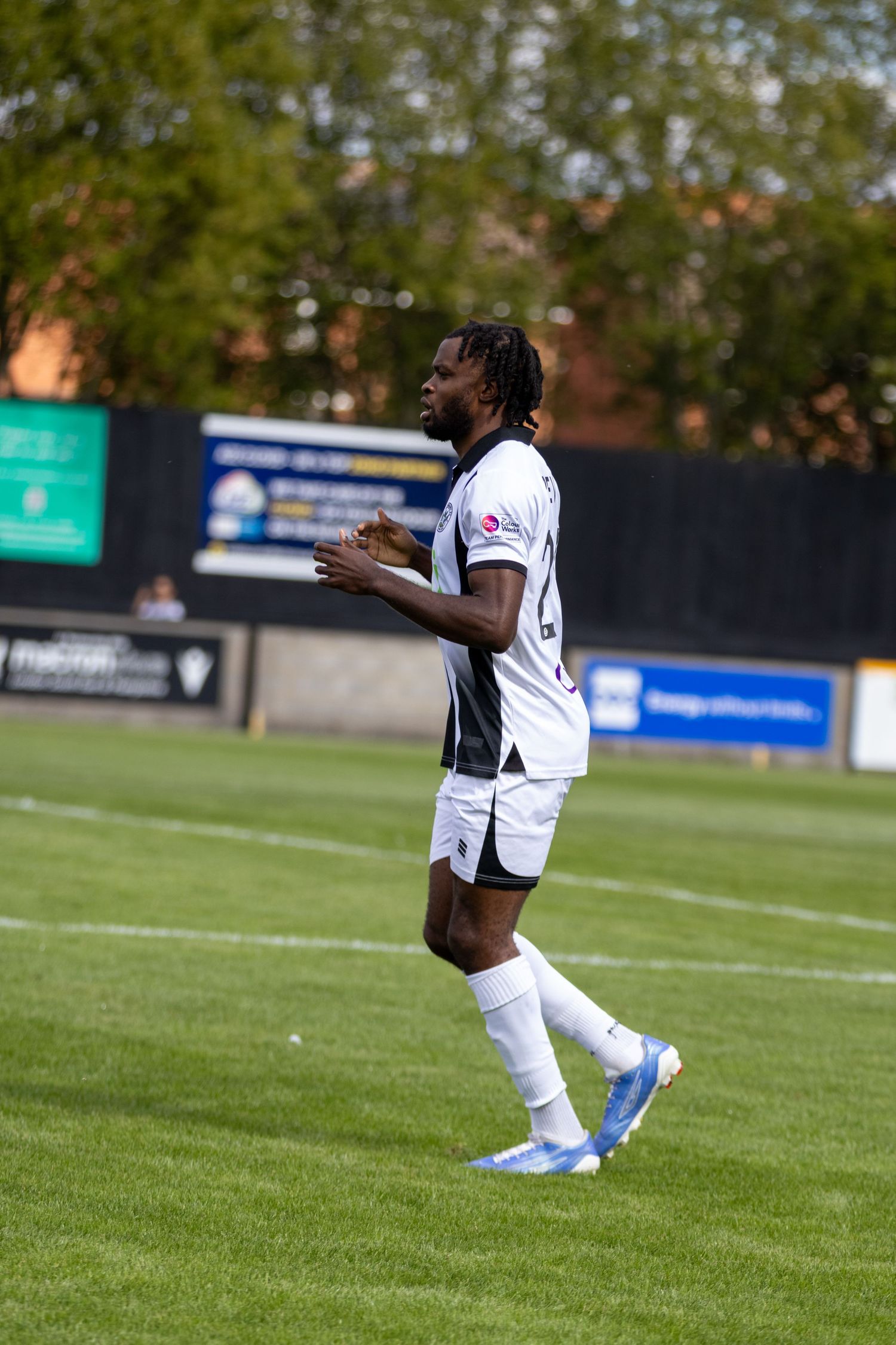 A soccer player in white and black kit with blue cleats walks on a green pitch during a match.