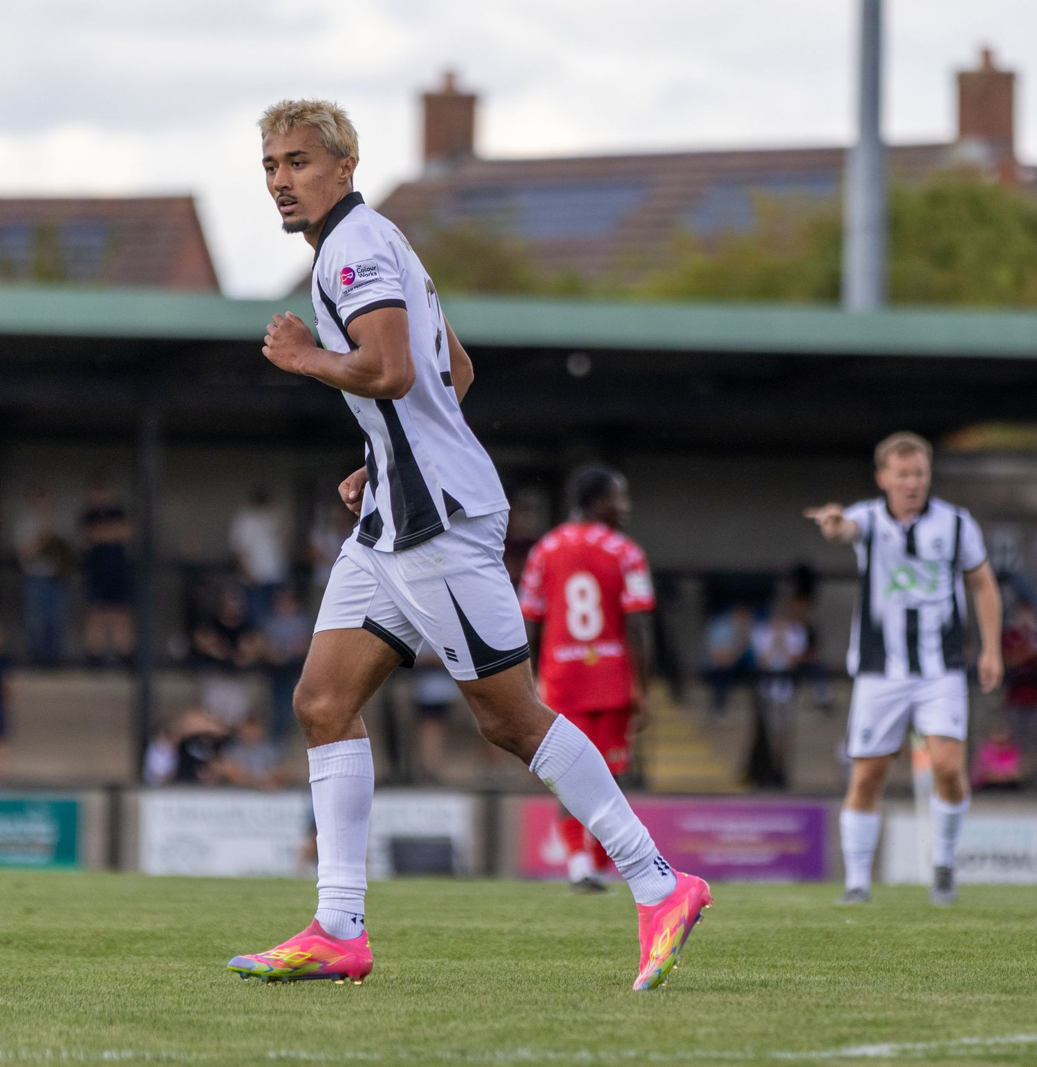 A footballer in black and white strip runs during a non-league match with red-kitted players in background.