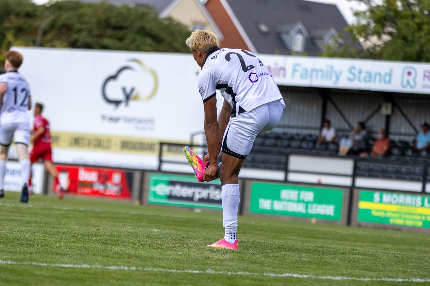 A footballer wearing white kit and pink boots bends down on the pitch during a non-league football game.