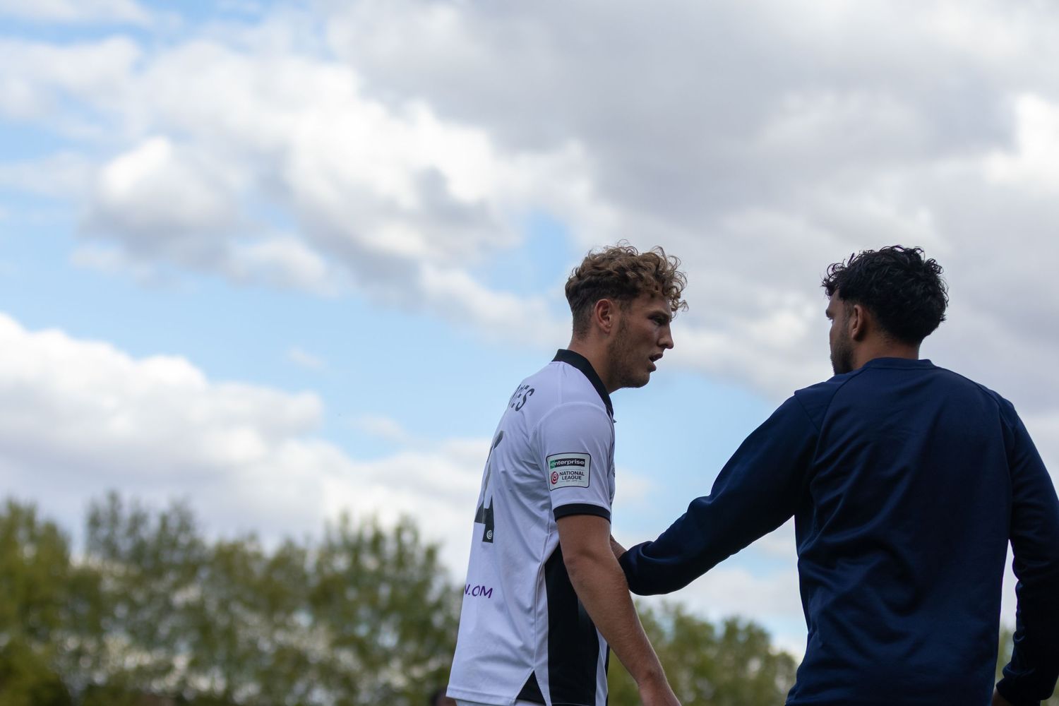 Two soccer players in white and navy uniforms having a discussion on a field with cloudy skies in the background.