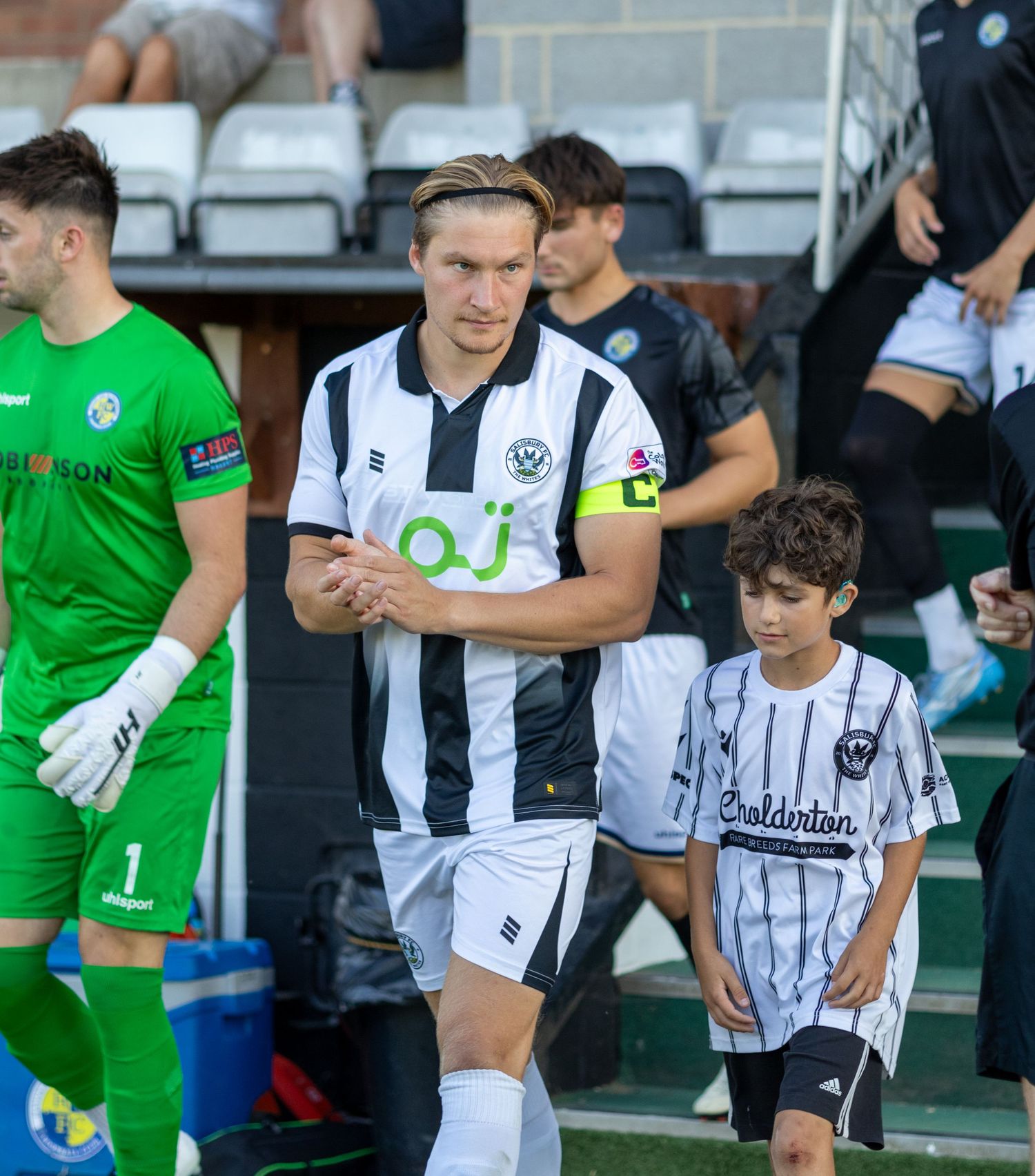 A footballer in black and white stripes walks onto the pitch with a young mascot during a match day.