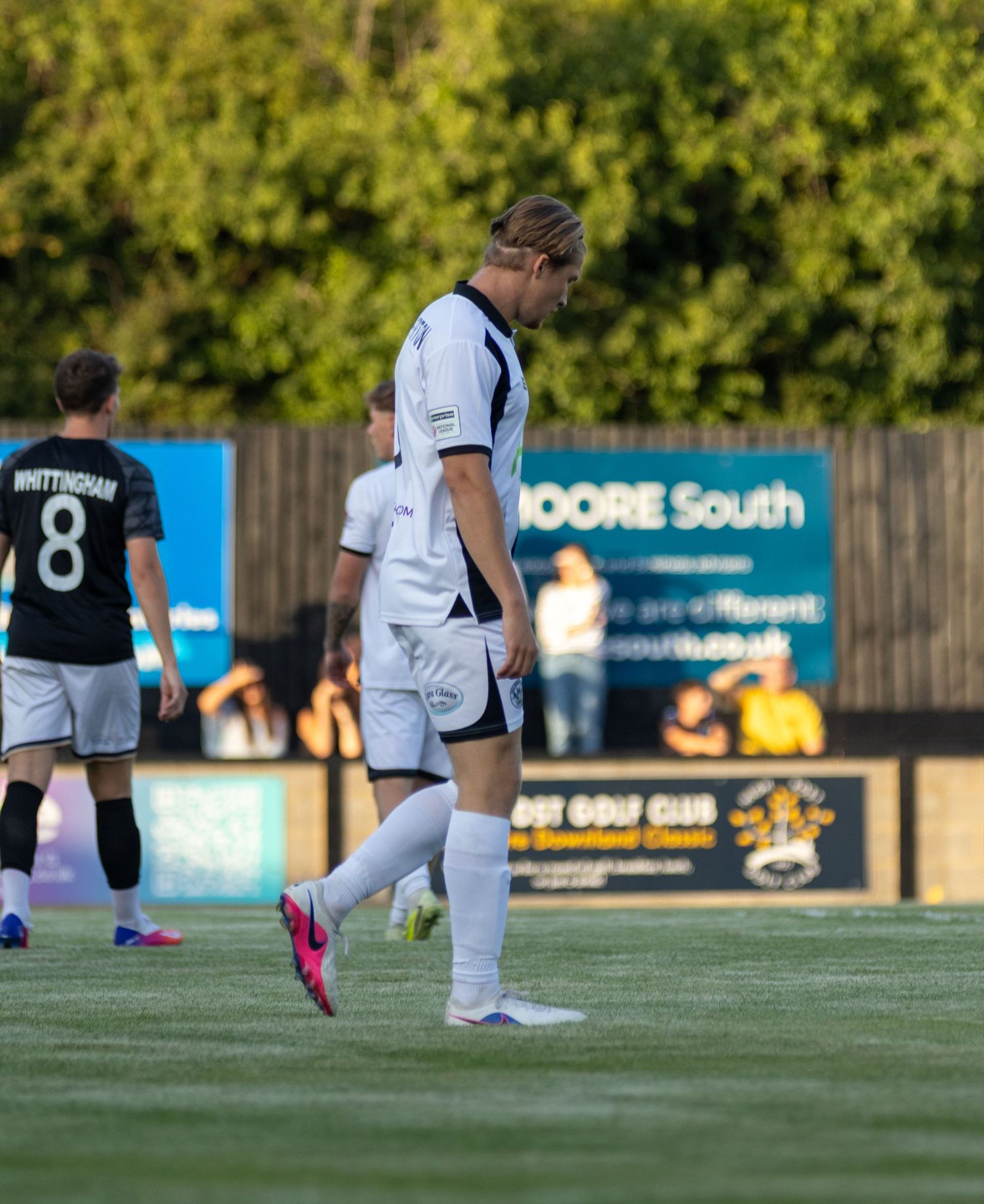 A blonde footballer in white kit walks across the pitch with Whittingham number 8 visible in the background.