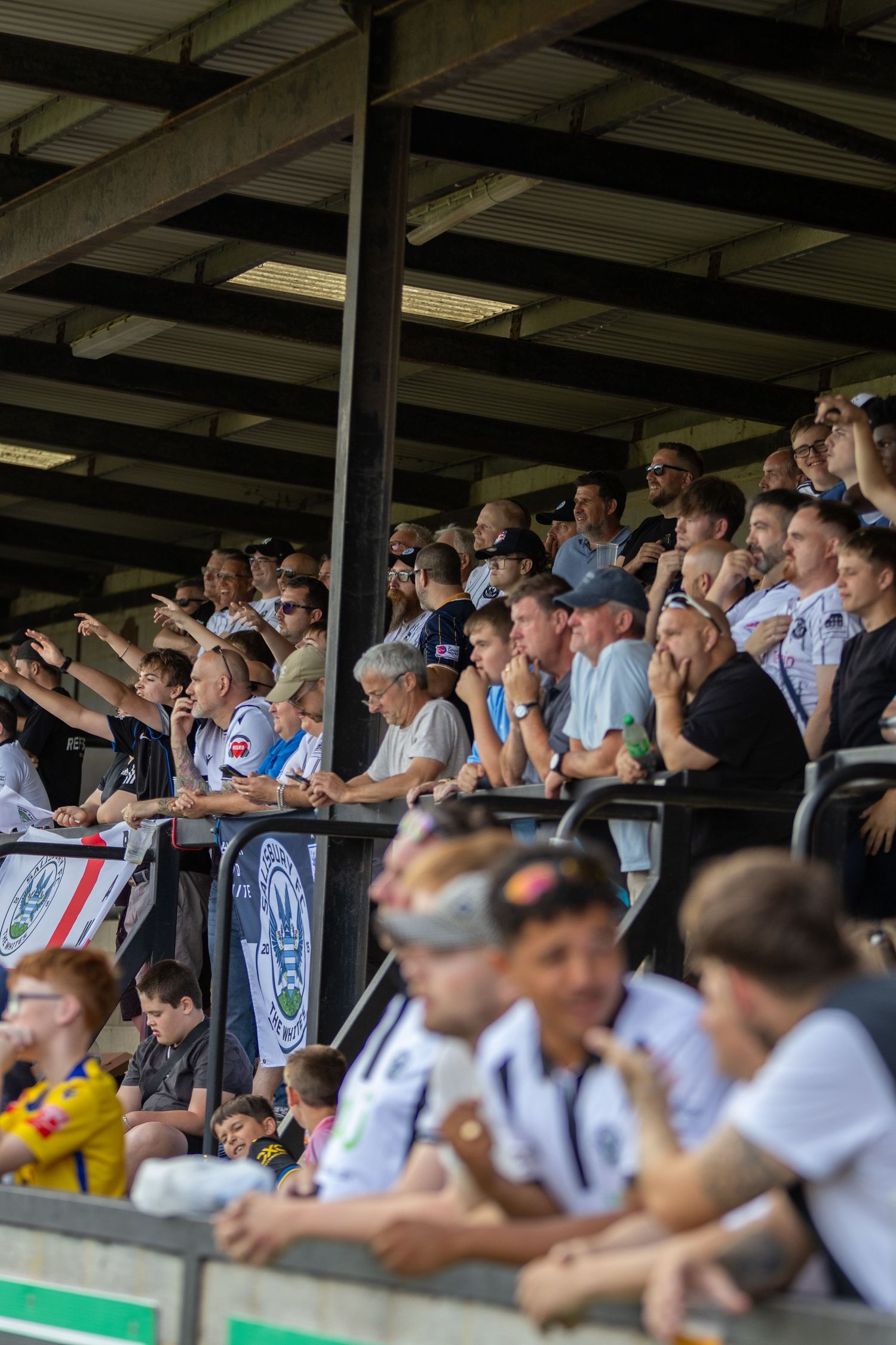 Football supporters watch intently from a crowded covered grandstand during a sunny afternoon match.