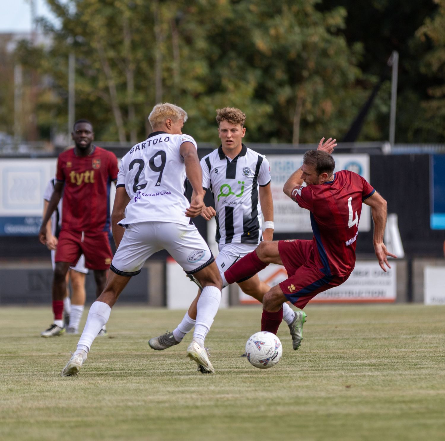 Two white-kit players challenge opponent number 4 who slides for the ball in sunny football match.