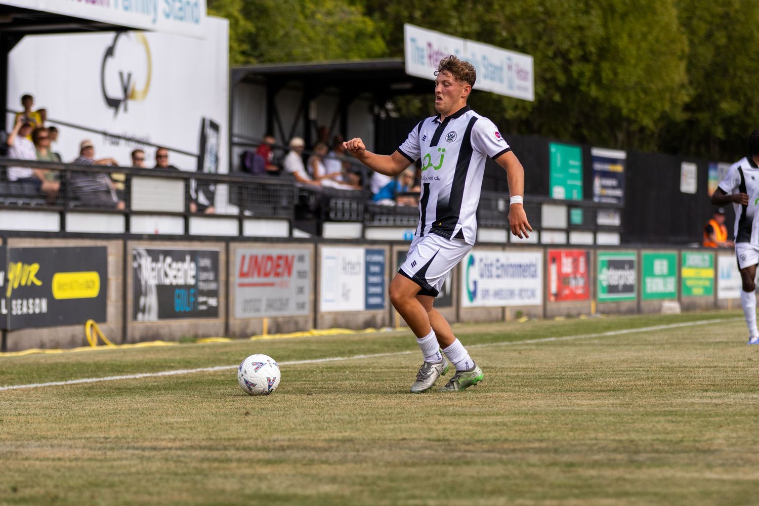 A soccer player in black and white kit runs with the ball on a dry grass pitch during a competitive outdoor match.