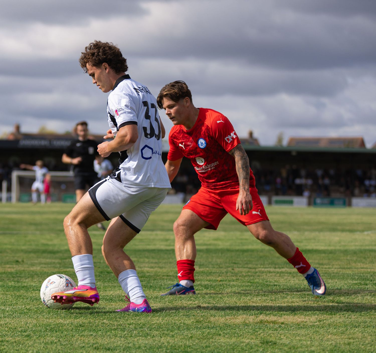 Two soccer players compete for the ball on a green pitch, one in white kit and one in red, during an outdoor match.