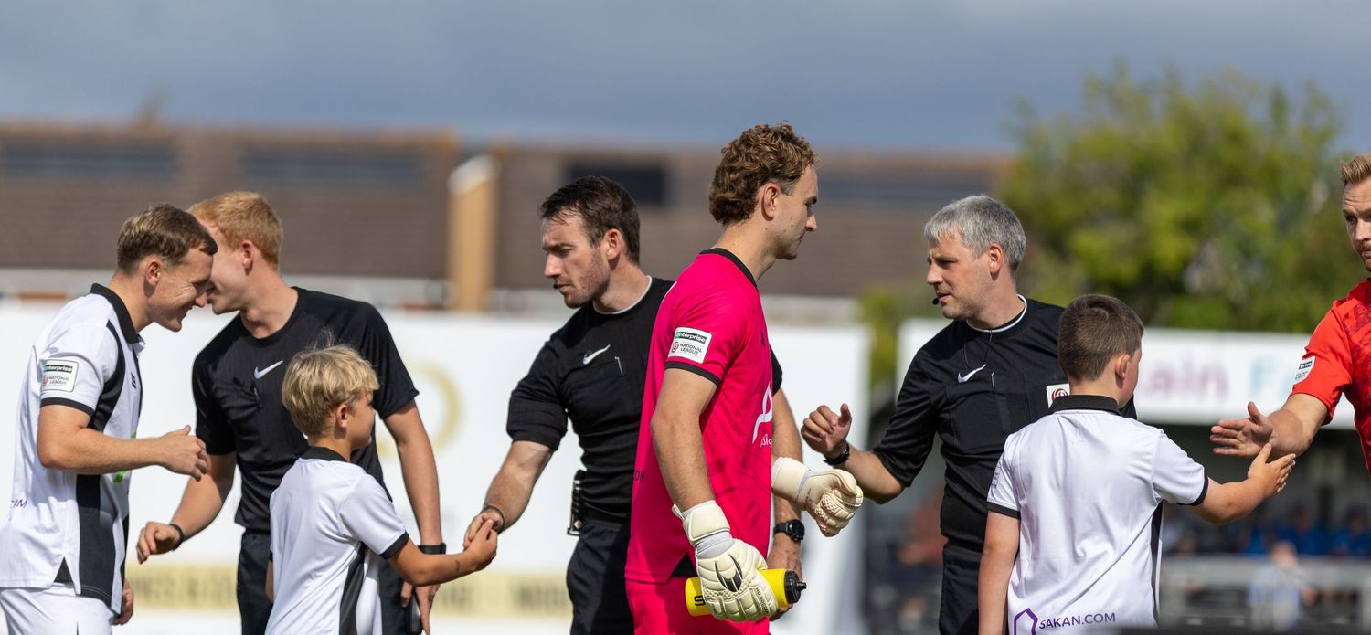 Football players shaking hands with young mascots before a match on a sunny day at a local football ground.
