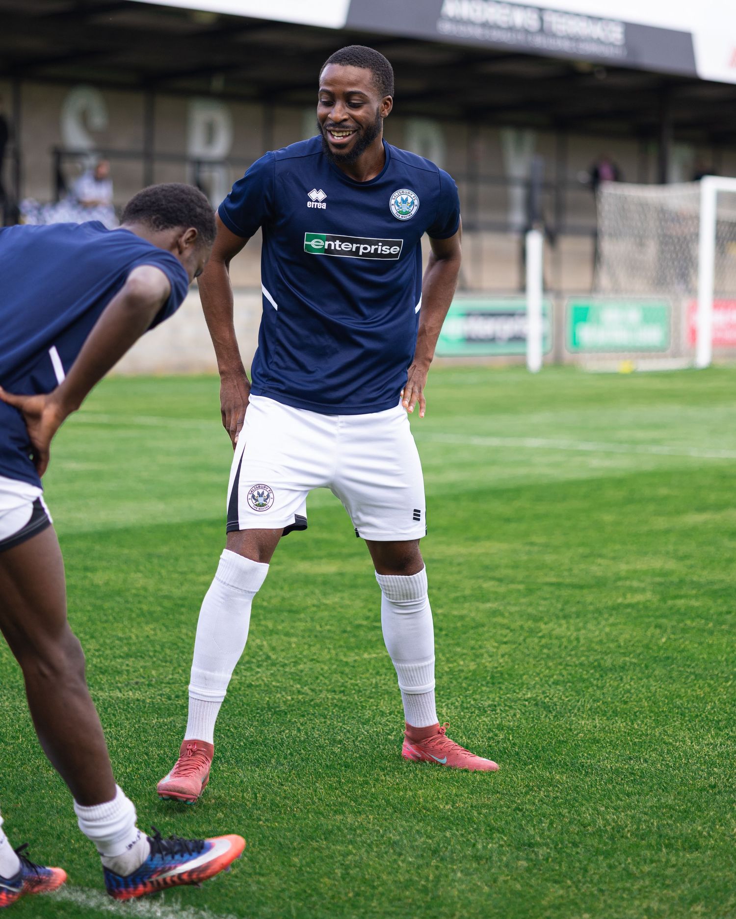 Two football players in navy Errea kits with Enterprise sponsor warm up on a grass pitch before a match.