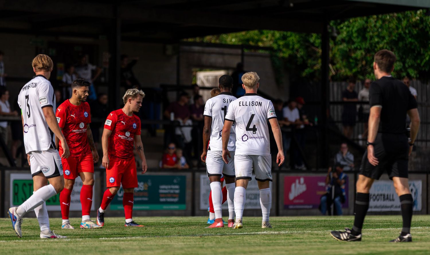 Football players in red and white kits face opponents during a sunny outdoor match near the penalty area.