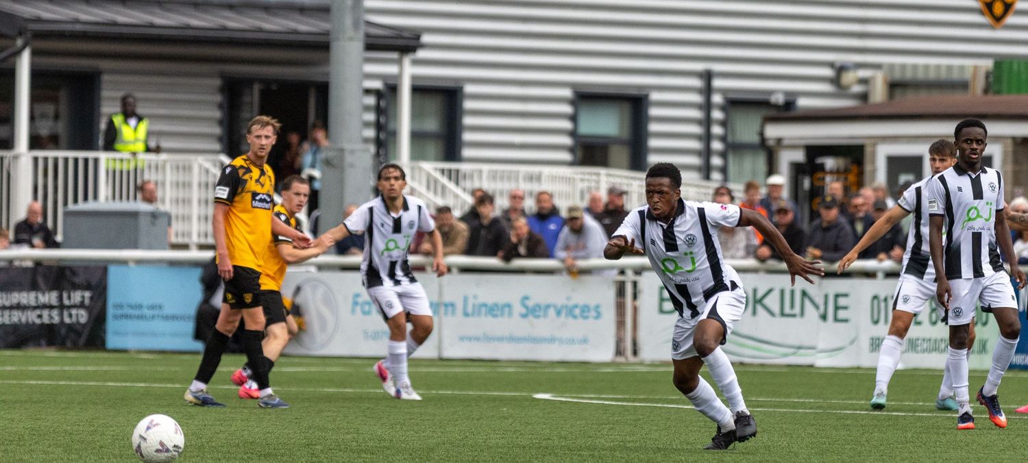 Soccer players compete on a green field during a match, with spectators and sponsor banners visible in the background.
