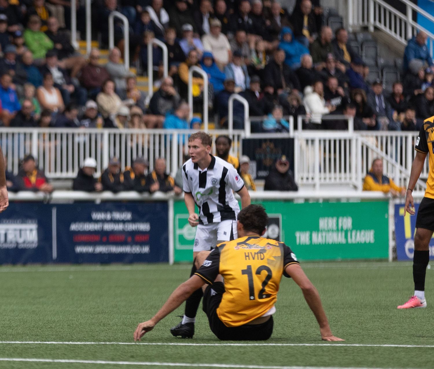 Soccer players in black and white vs yellow uniforms compete on a green field with spectators in the stands.